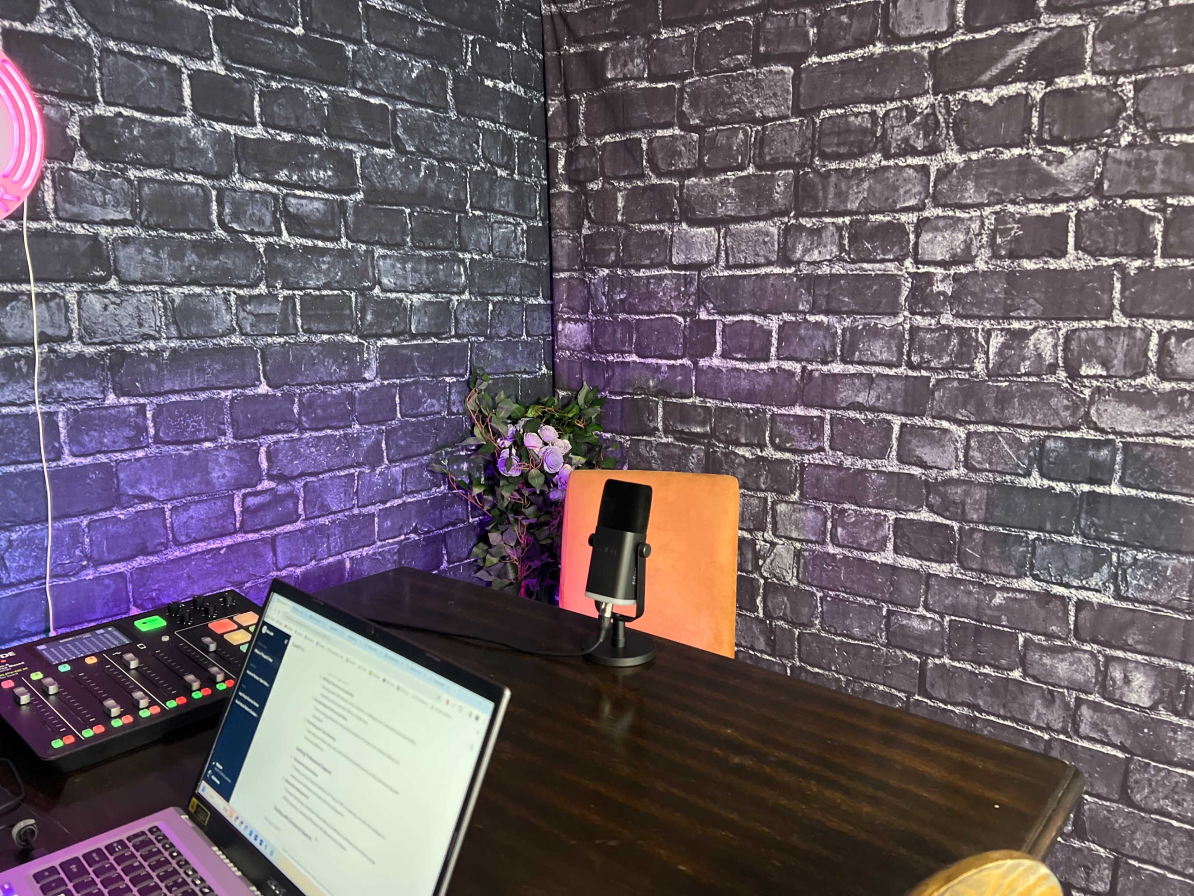 A workspace with a microphone on a table, a laptop open, and a corner wall featuring brick texture, complemented by some greenery.