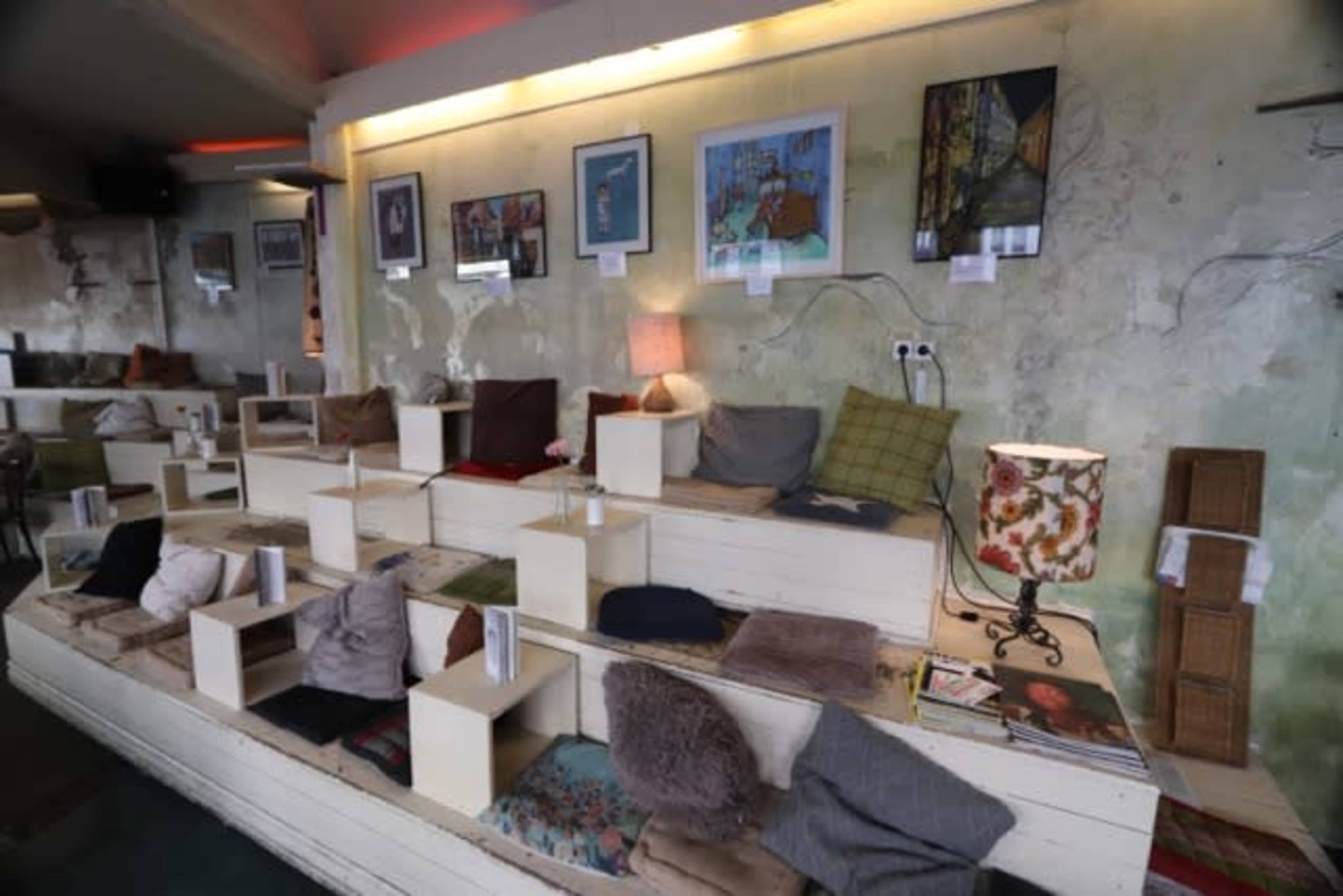 The image shows a cozy interior space with tiered seating, various cushions, and framed artworks displayed on the walls.