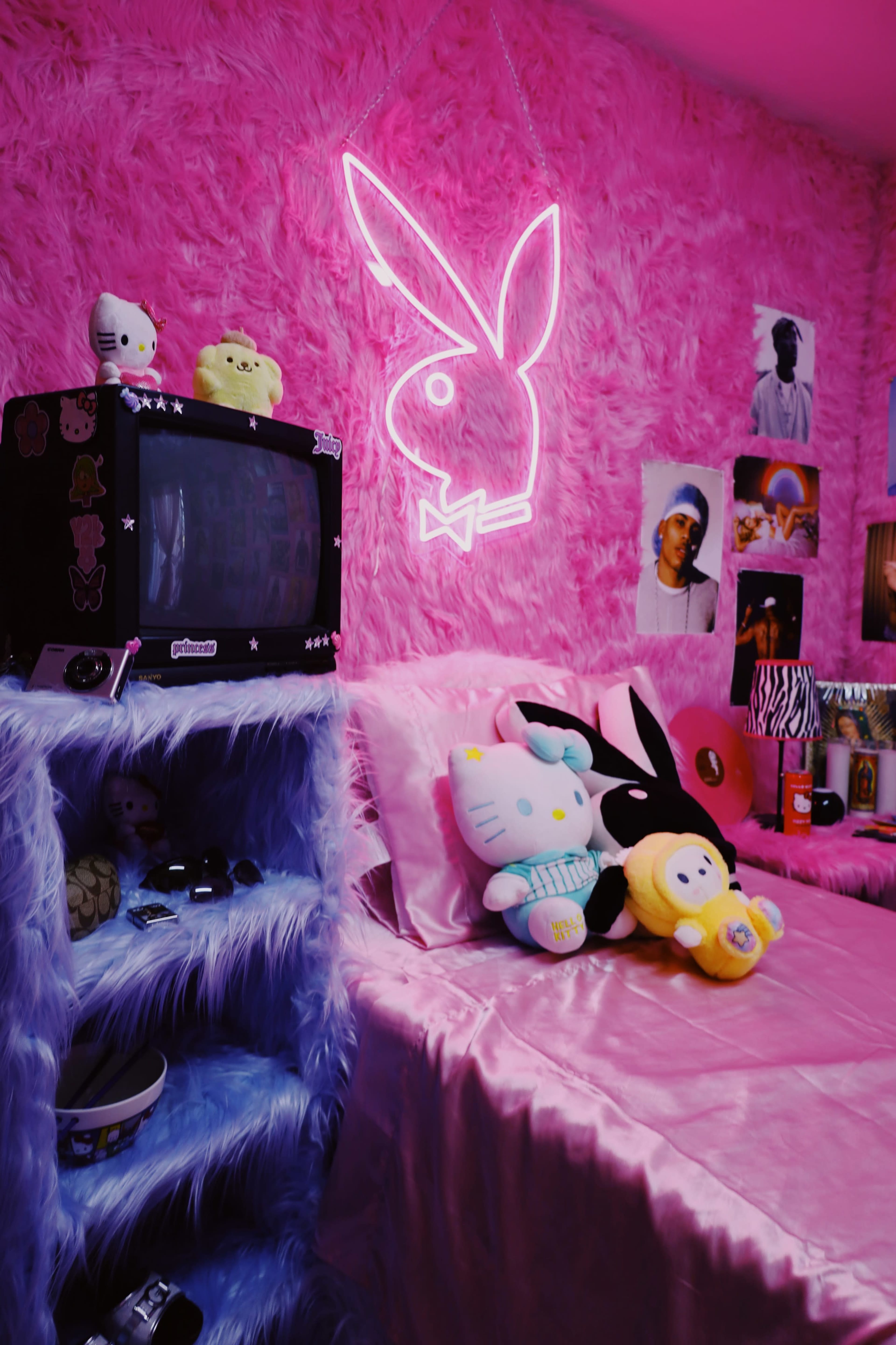 The image shows a colorful bedroom with pink fluffy walls, a bed covered in a pink satin blanket, and various decorative pillows, including plush toys, alongside a retro television and wall art featuring celebrities.