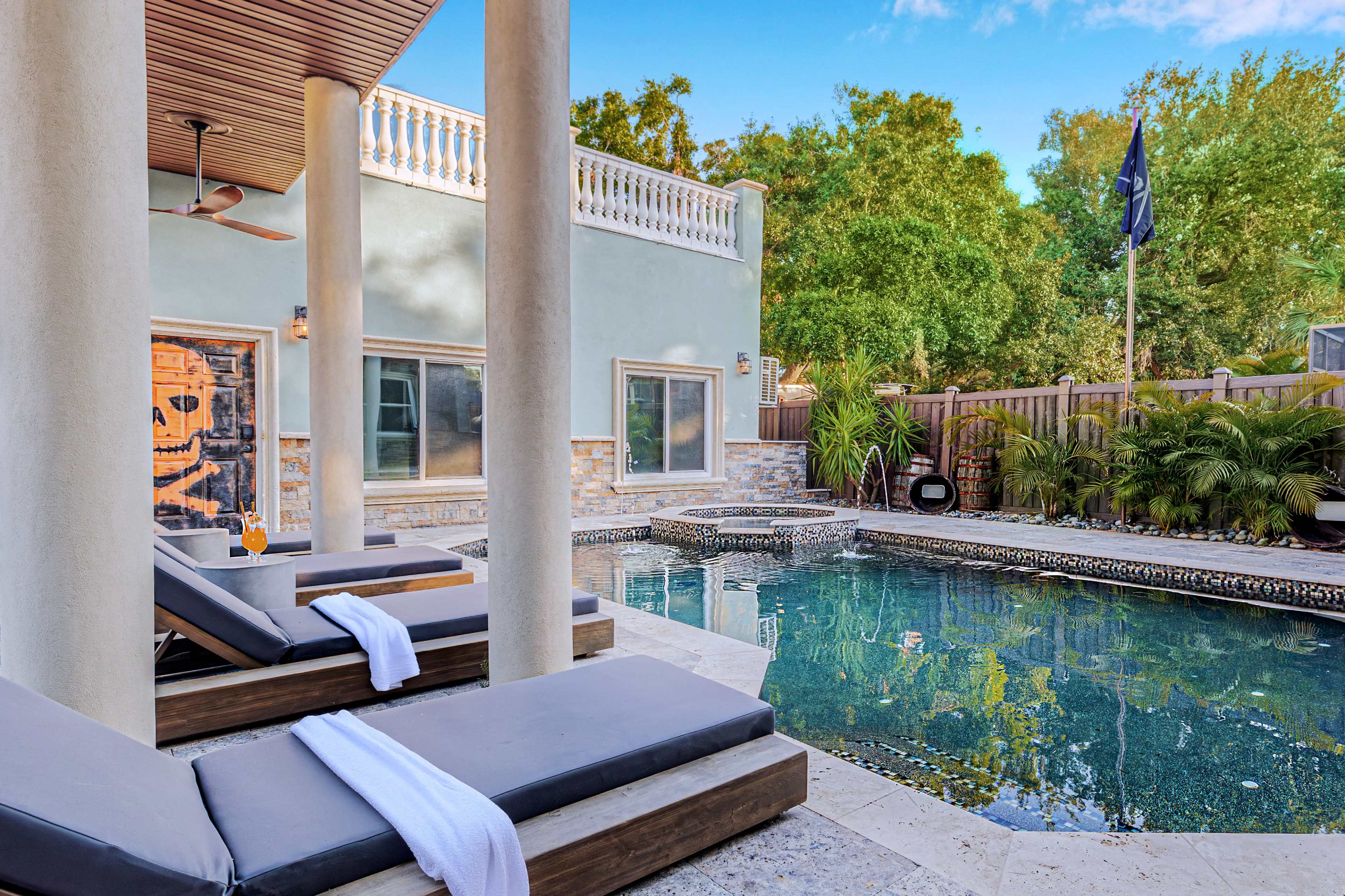 The image shows a modern outdoor pool area with lounge chairs, surrounded by greenery and a stone fence.