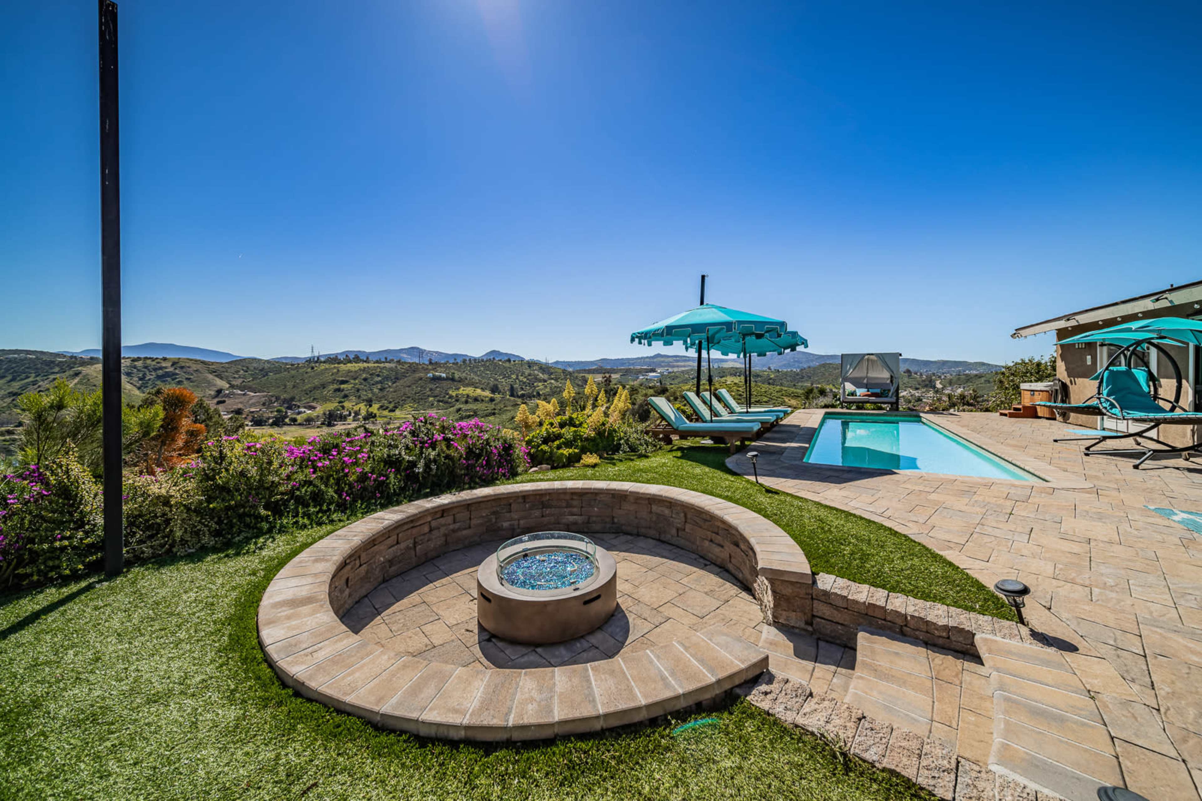 A landscaped backyard with a spa, swimming pool, and lounge chairs, surrounded by lush greenery and mountains in the distance.