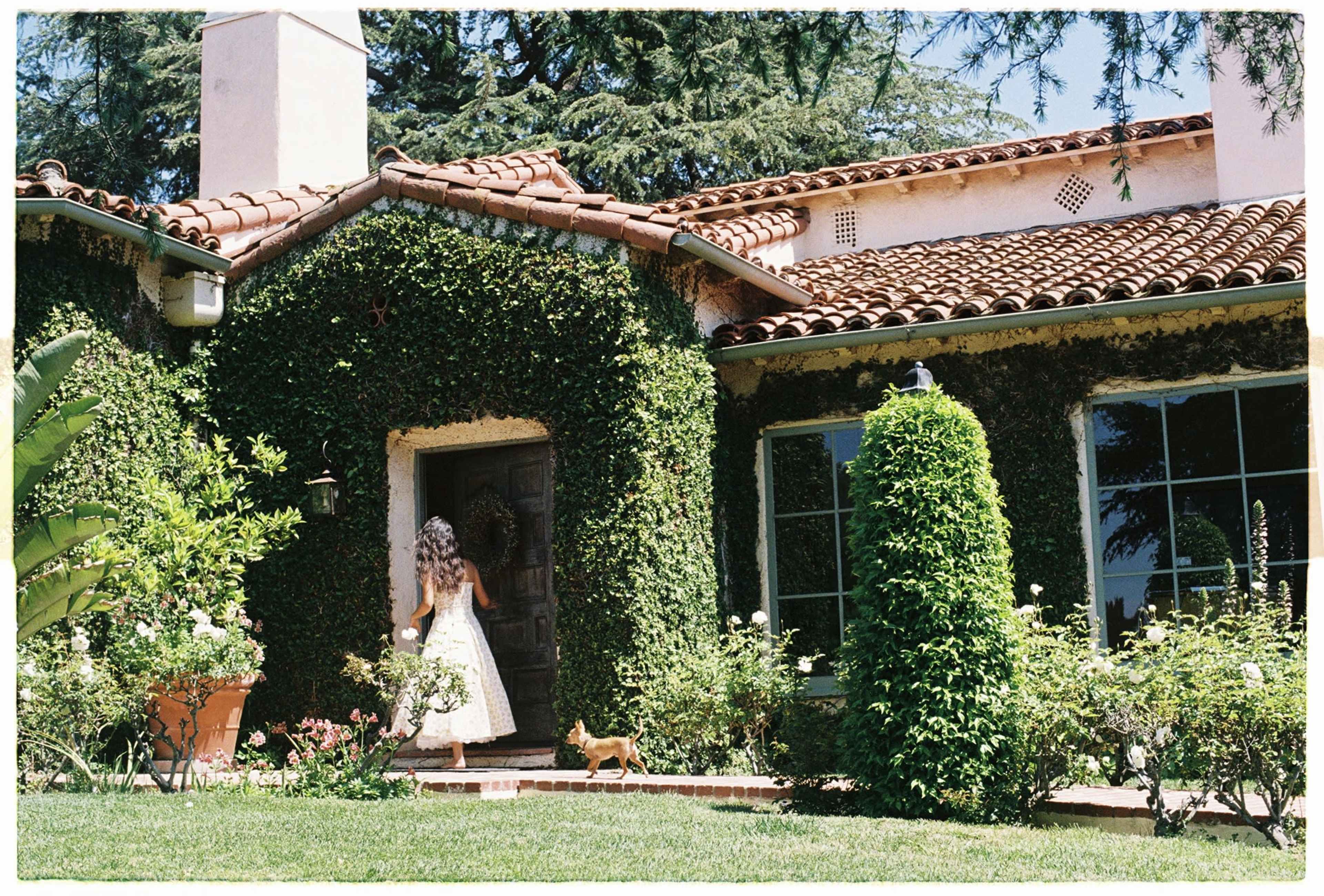 A girl wearing a white dress walks toward a house covered in ivy, with a small dog following her.