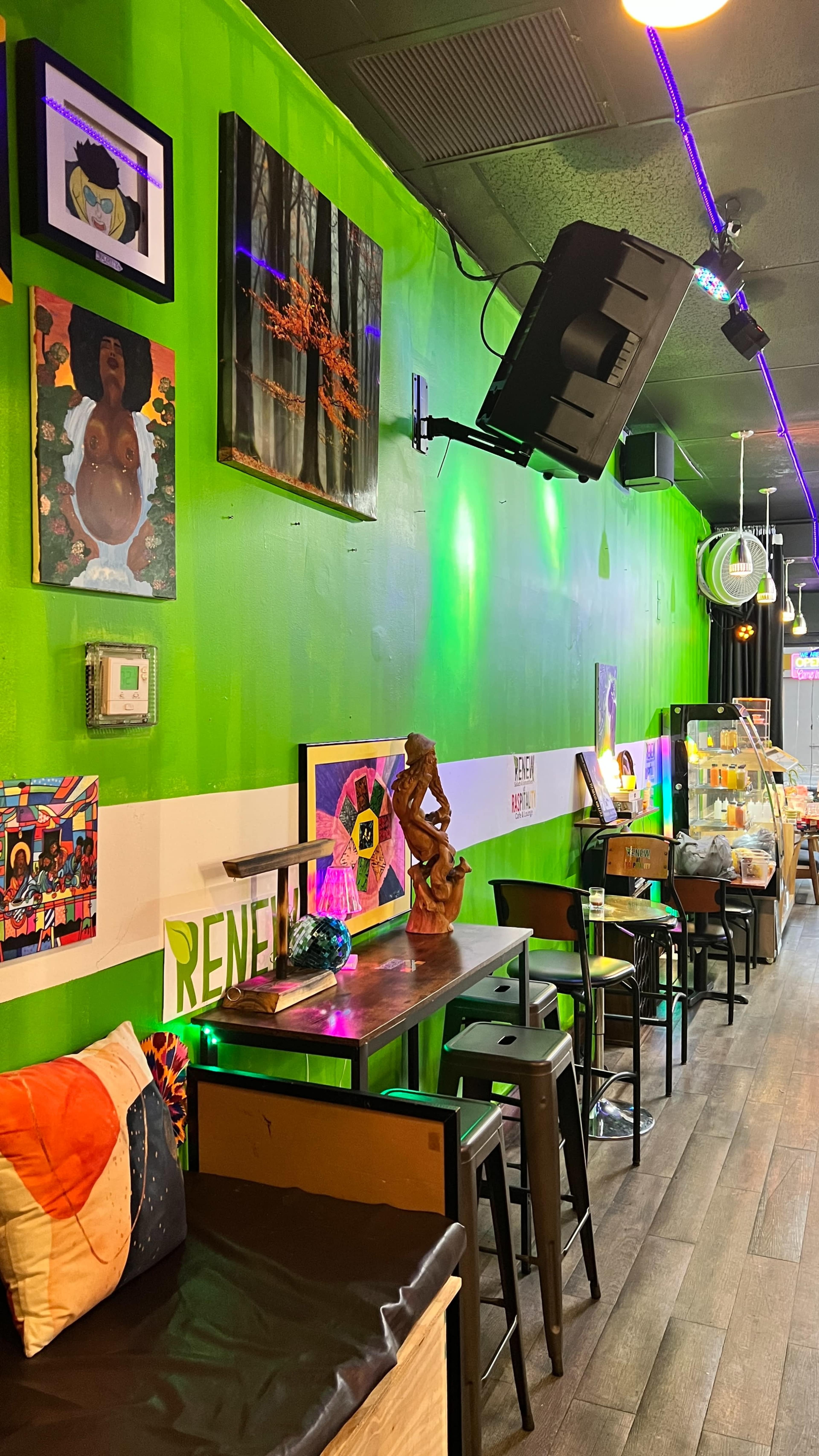 The image shows a vibrant café interior featuring colorful artwork on green walls, wooden stools, and decorative items displayed on shelves.