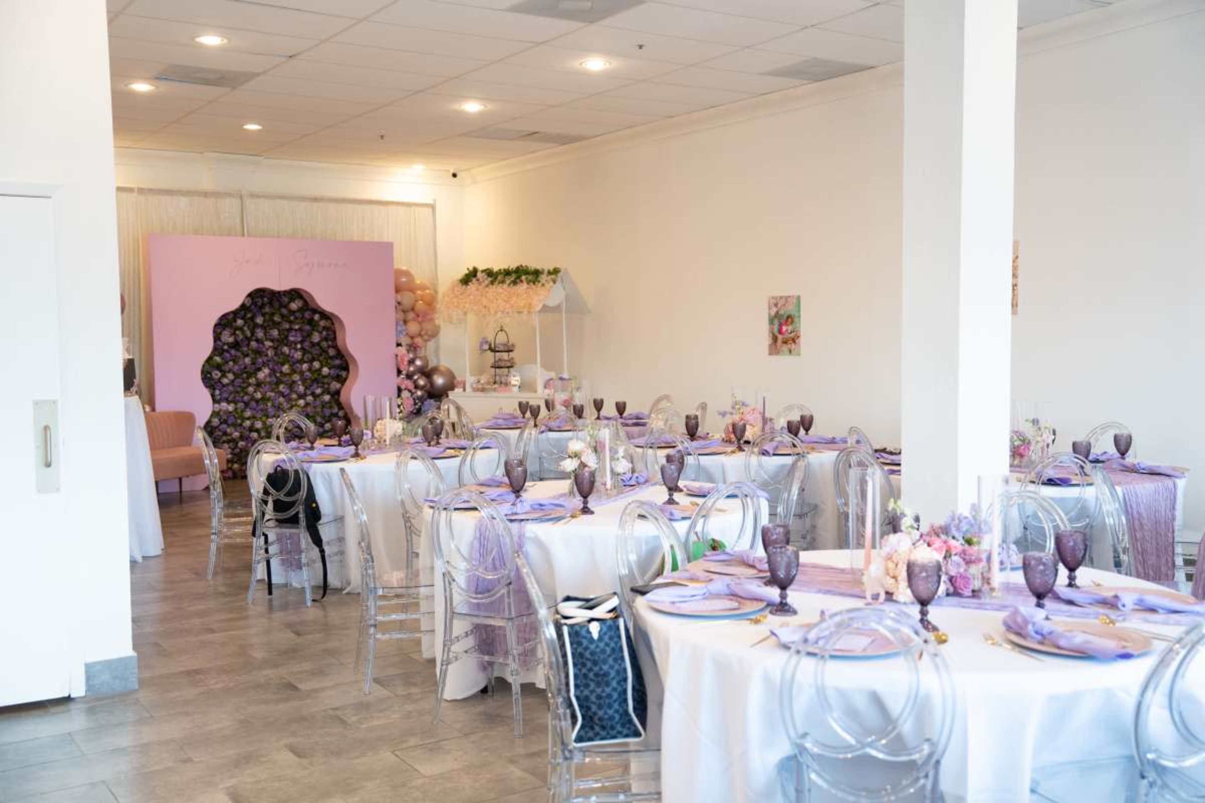 The image shows a decorated event space with several tables set for a gathering, featuring pastel-colored tableware and a backdrop adorned with balloons and floral arrangements.