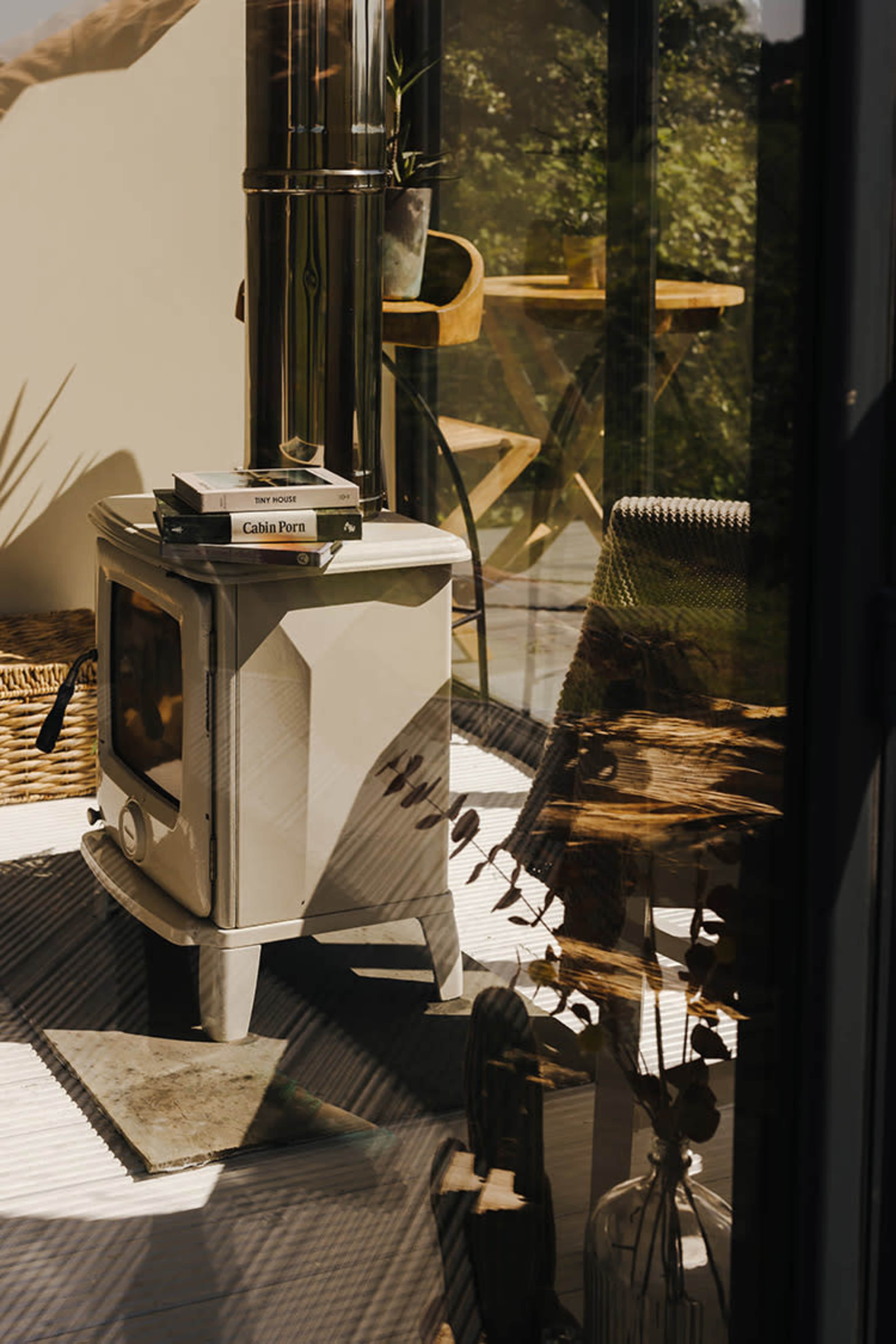 A small gray stove with books on top, surrounded by a cozy space with a woven basket, a chair, and plants, all reflected in glass.