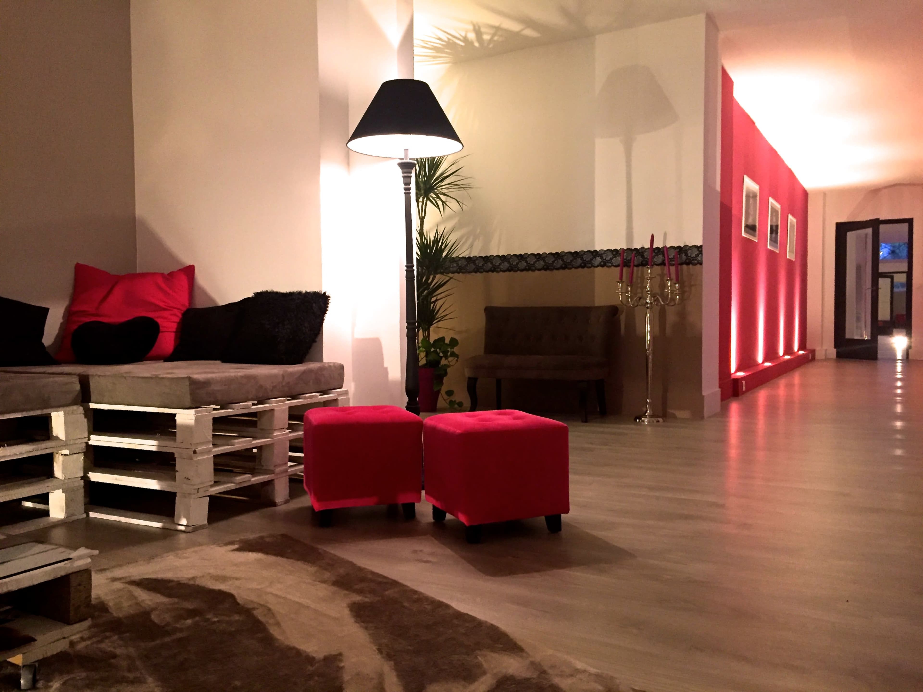 A modern interior space featuring a gray sofa made from pallets, two red ottomans, a lamp, and a corridor with pink walls illuminated by soft lighting.