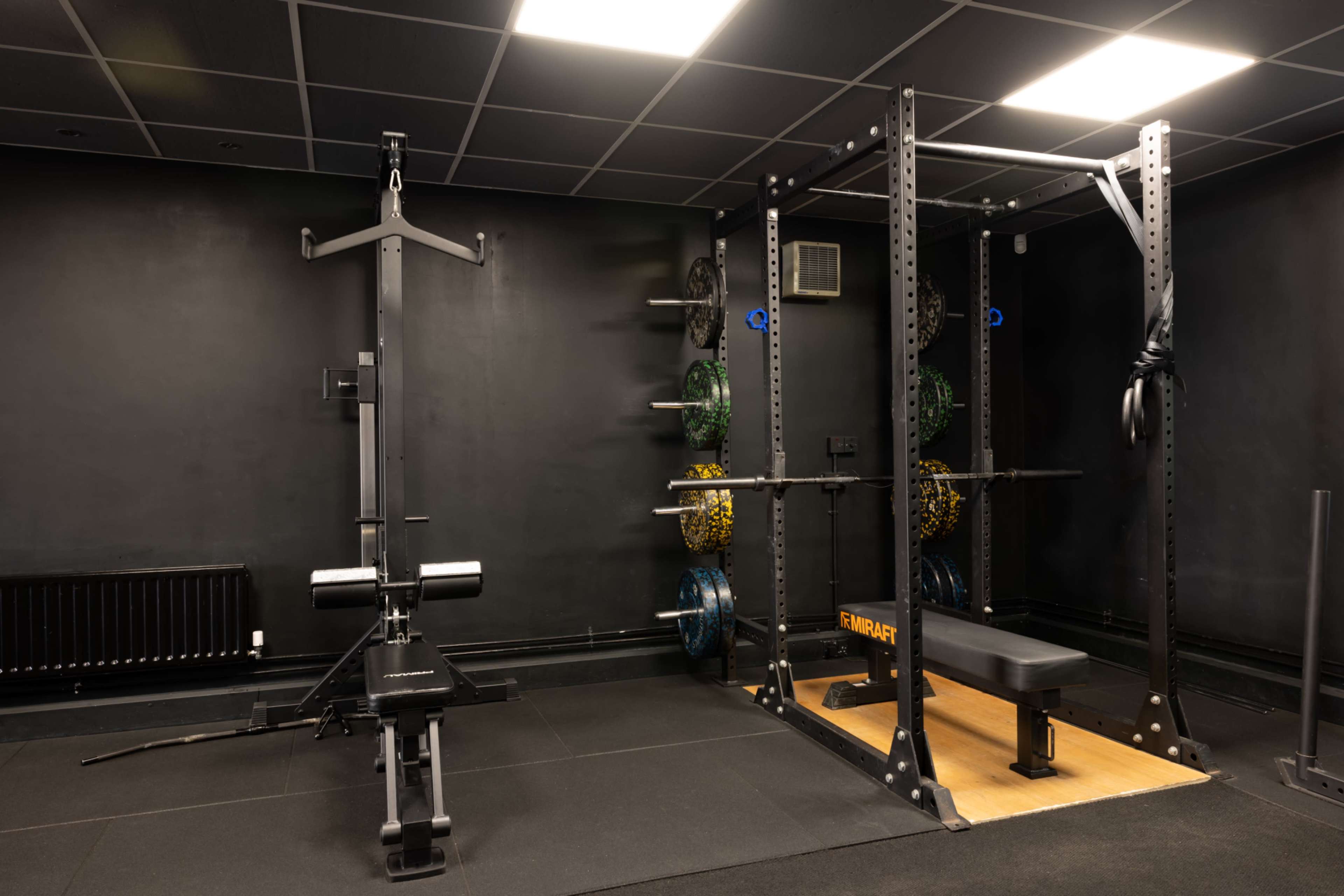 The image shows a black-walled gym interior featuring a weightlifting rack, a bench press, and various weight plates neatly arranged on a wall-mounted rack.