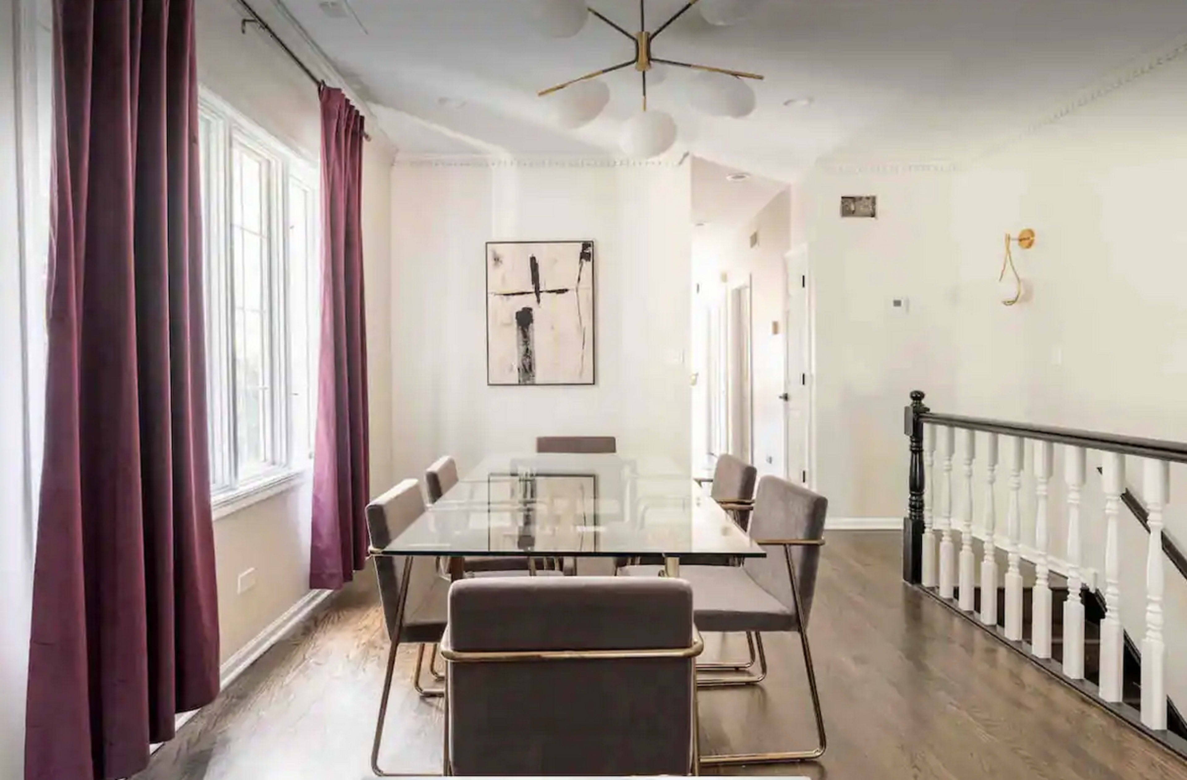 A modern dining room features a glass table surrounded by gray chairs, with large windows draped in burgundy curtains.
