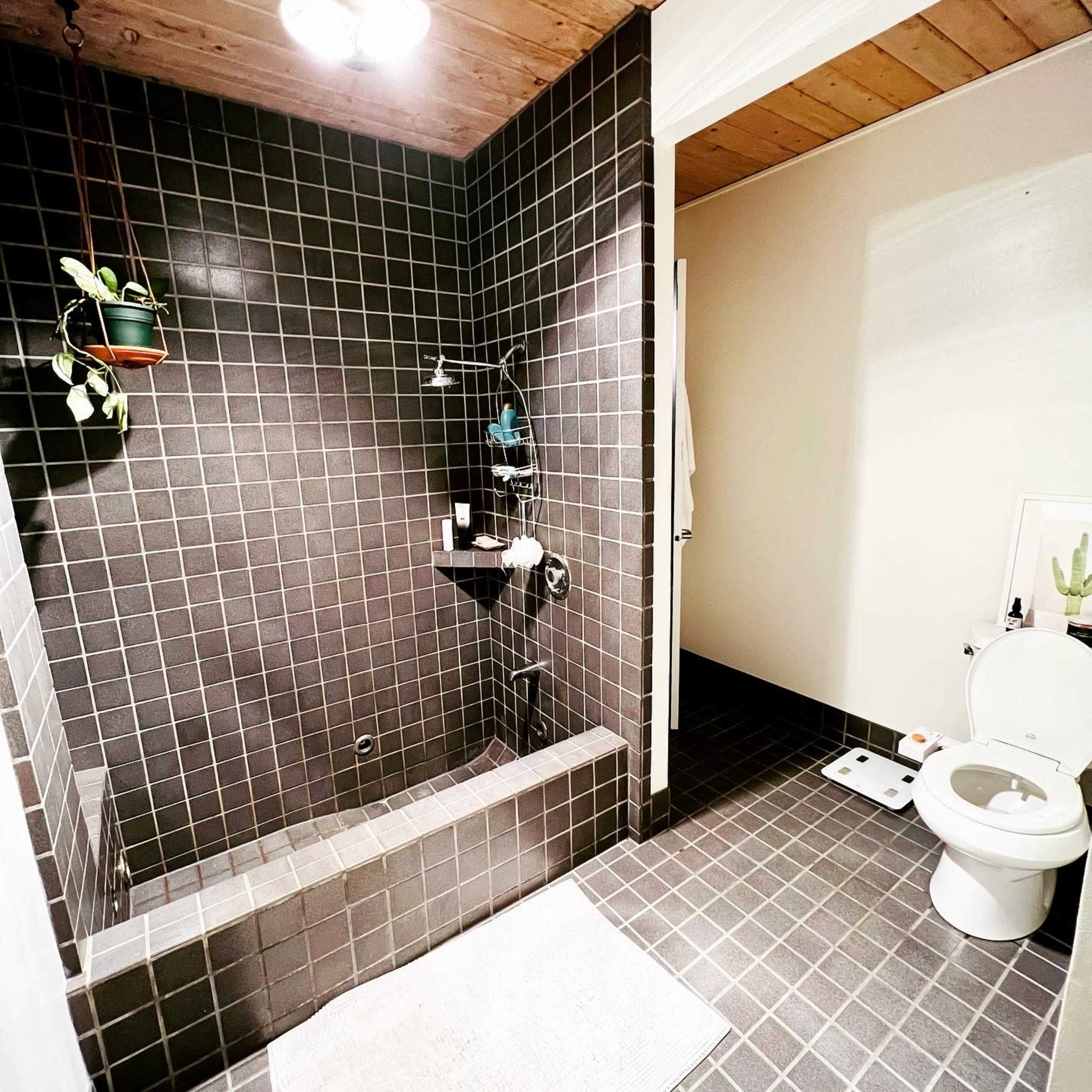 A dark tiled bathroom featuring a shower area with a bench and a separate toilet.