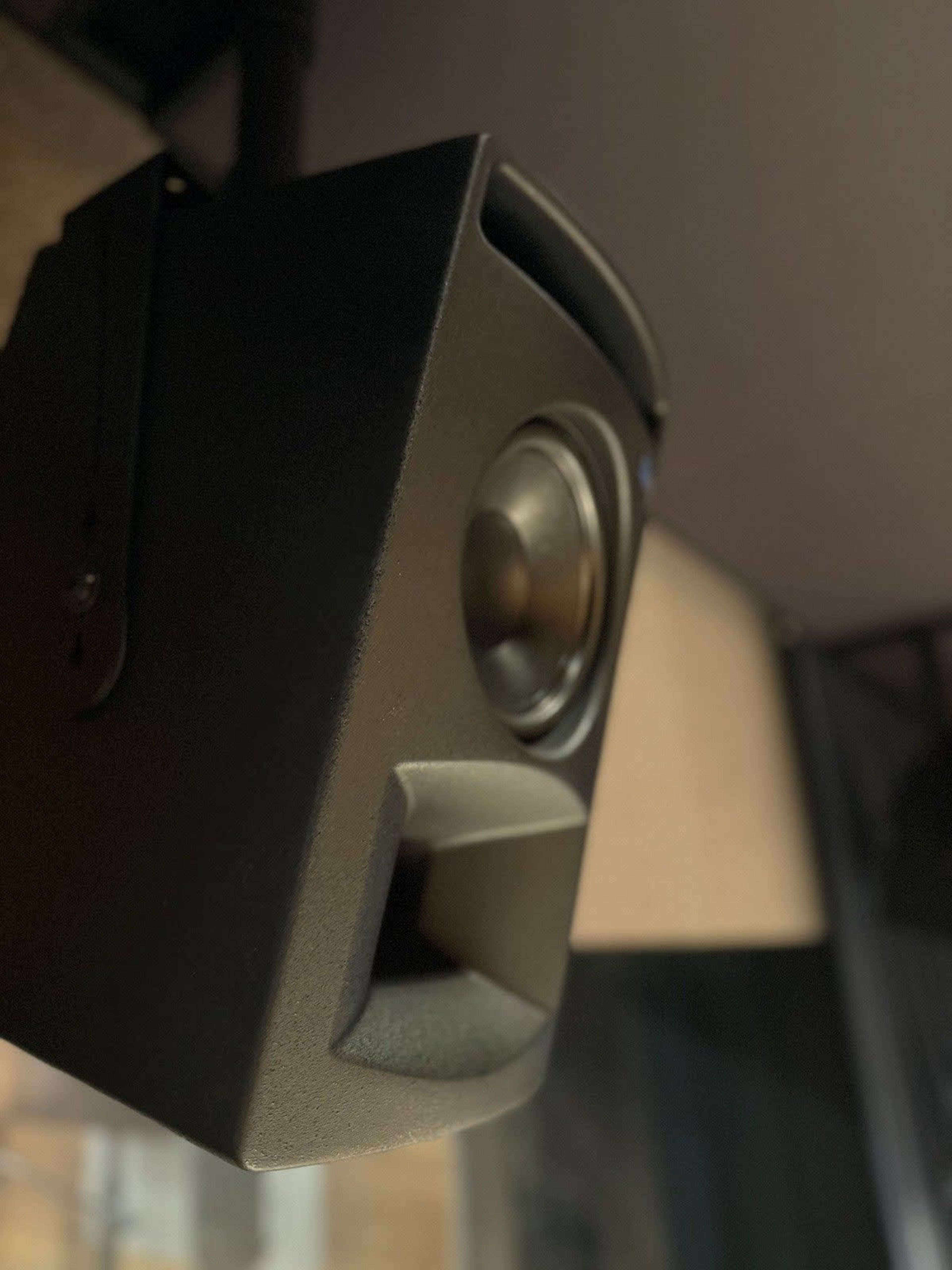 The image shows a black speaker mounted on a wall, angled slightly downward.