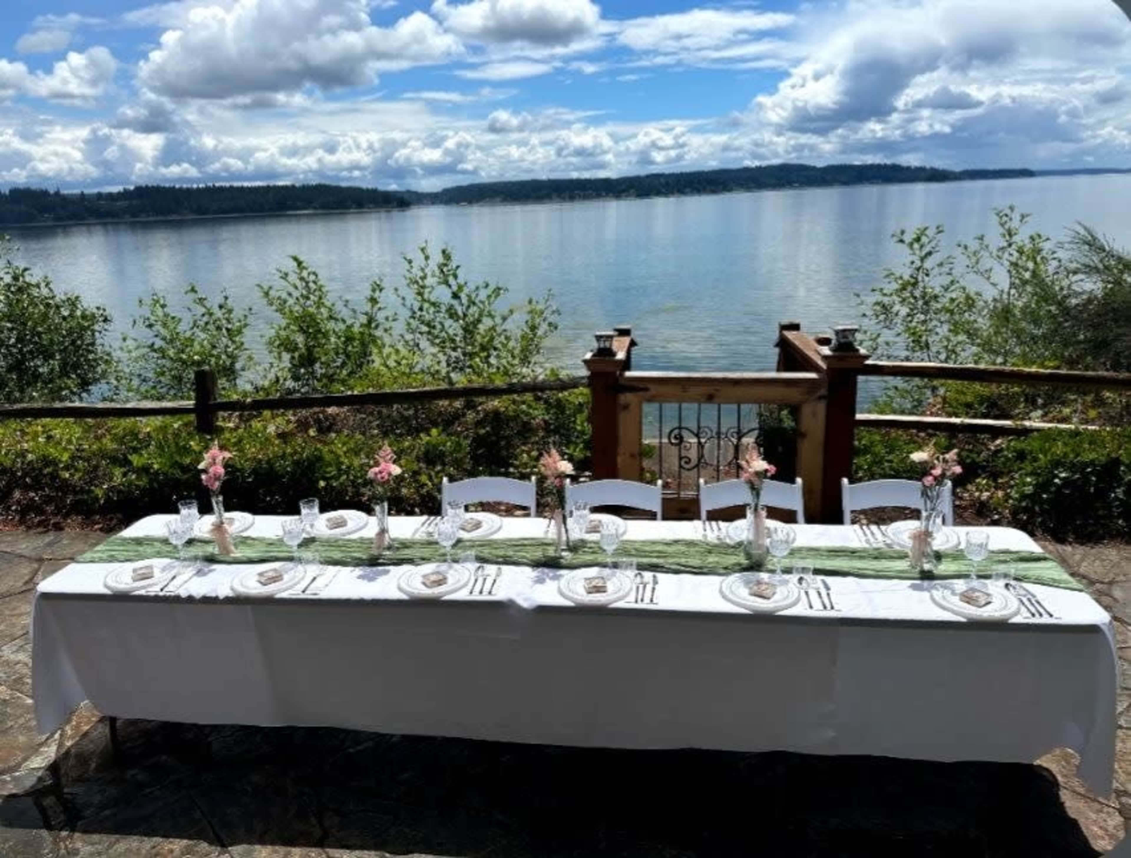 A long, elegantly set dining table with a white tablecloth is positioned by a scenic waterfront, surrounded by greenery under a cloudy sky.