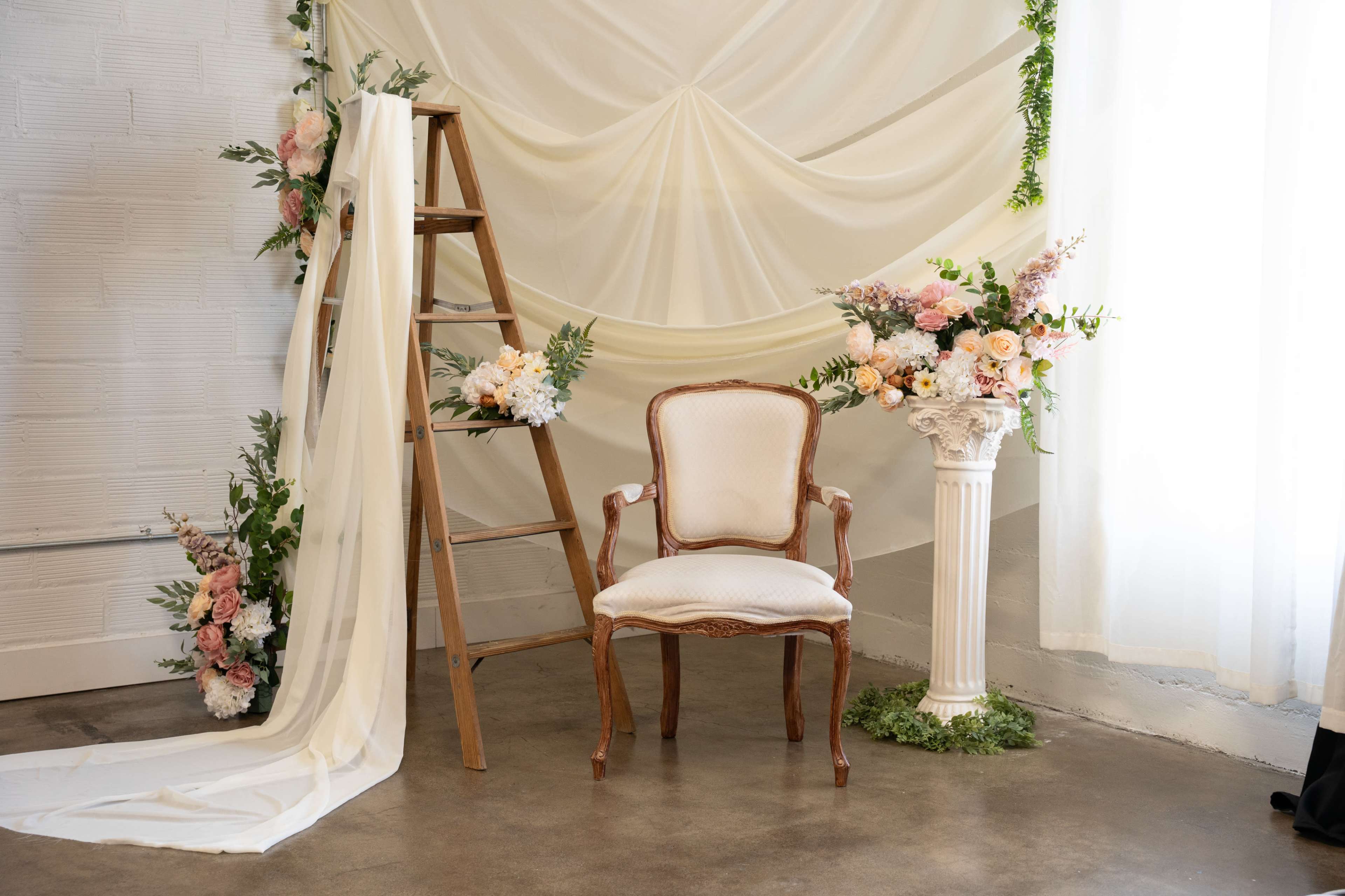 The image features a decorated corner with a white chair, a wooden ladder draped with fabric, and floral arrangements in vases, set against a backdrop of soft drapes.