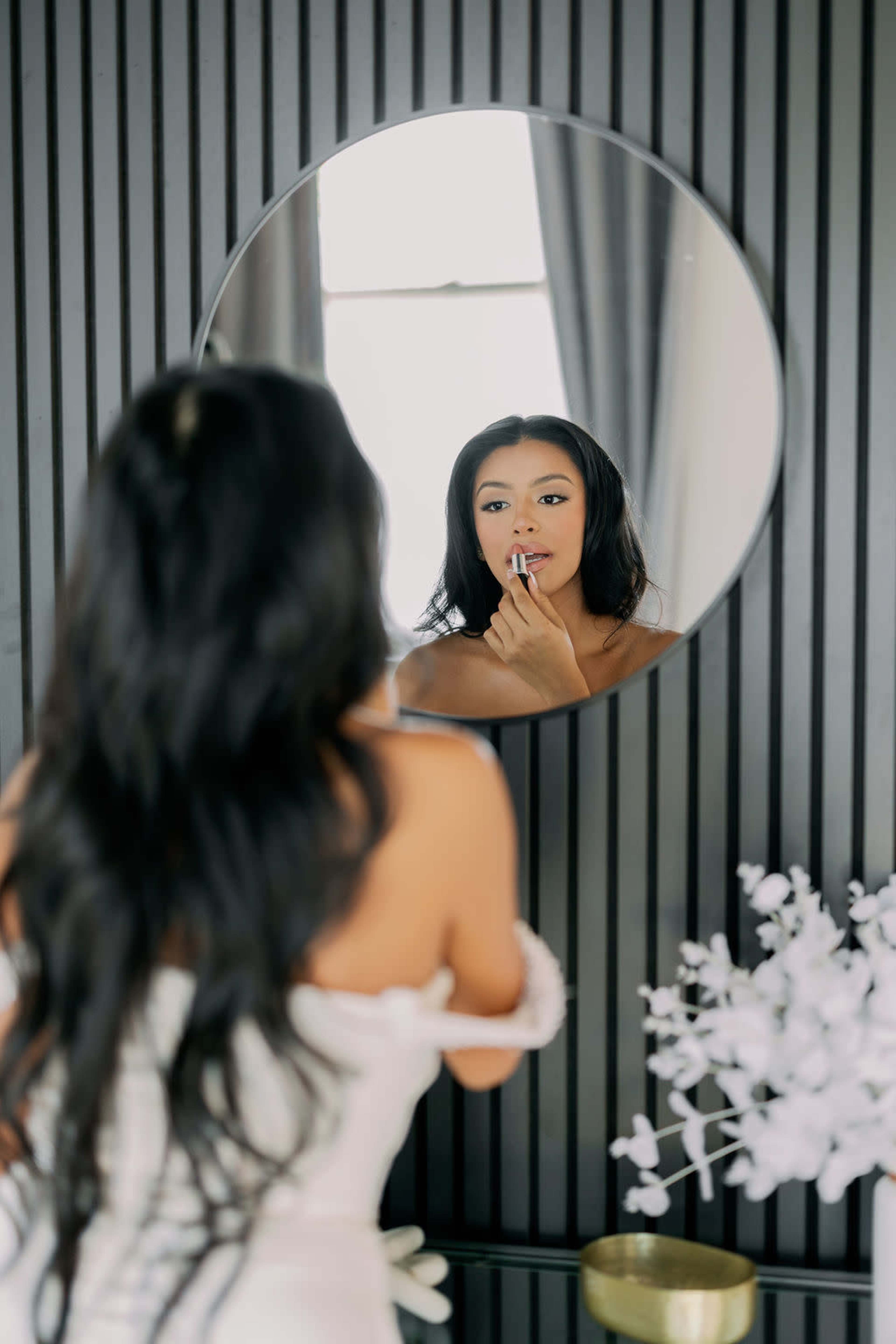 A woman with long hair applies makeup while looking into a round mirror mounted on a dark wood-paneled wall.