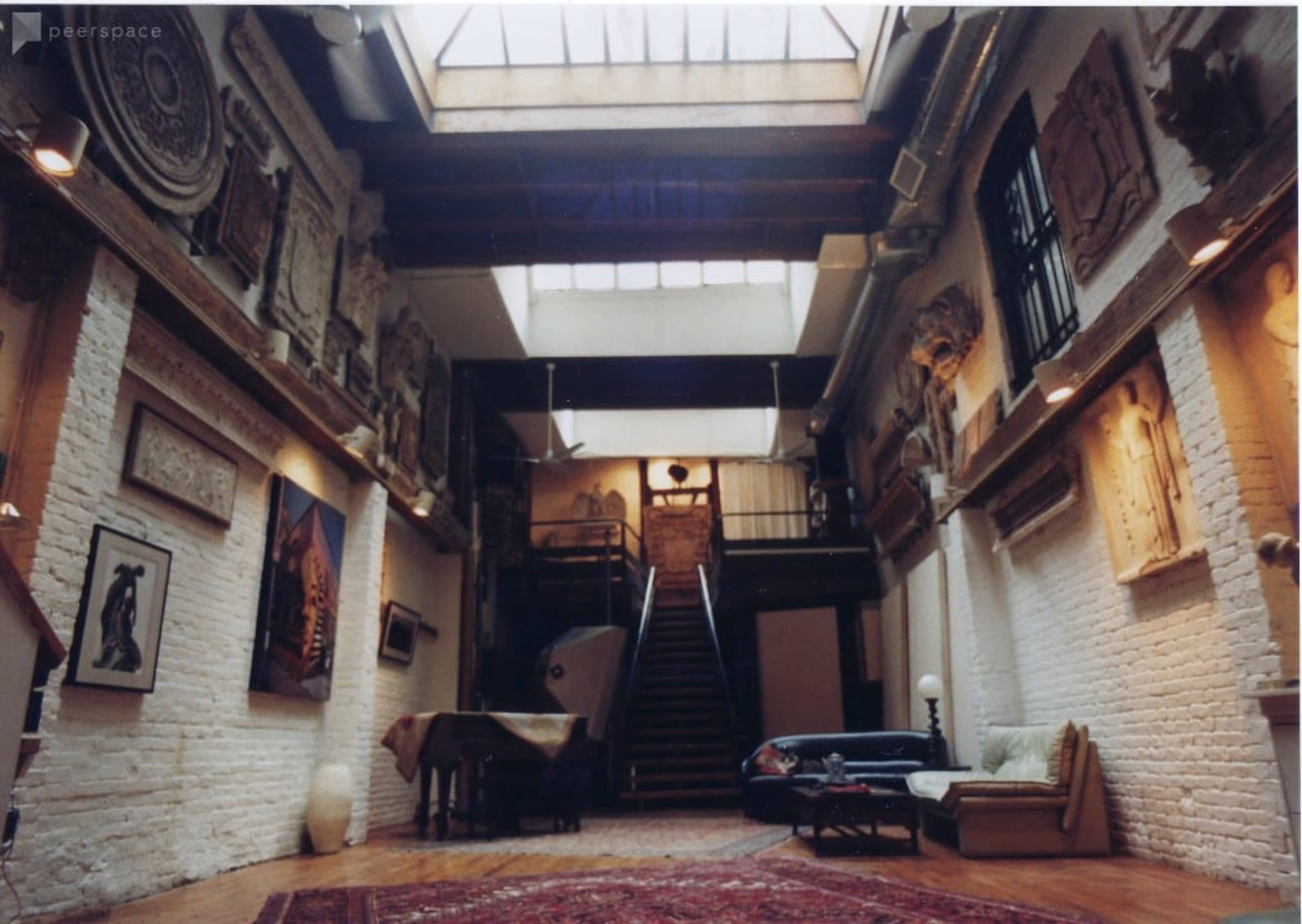 An art-filled loft interior with exposed brick walls, a wooden floor, and a staircase leading to an upper level.
