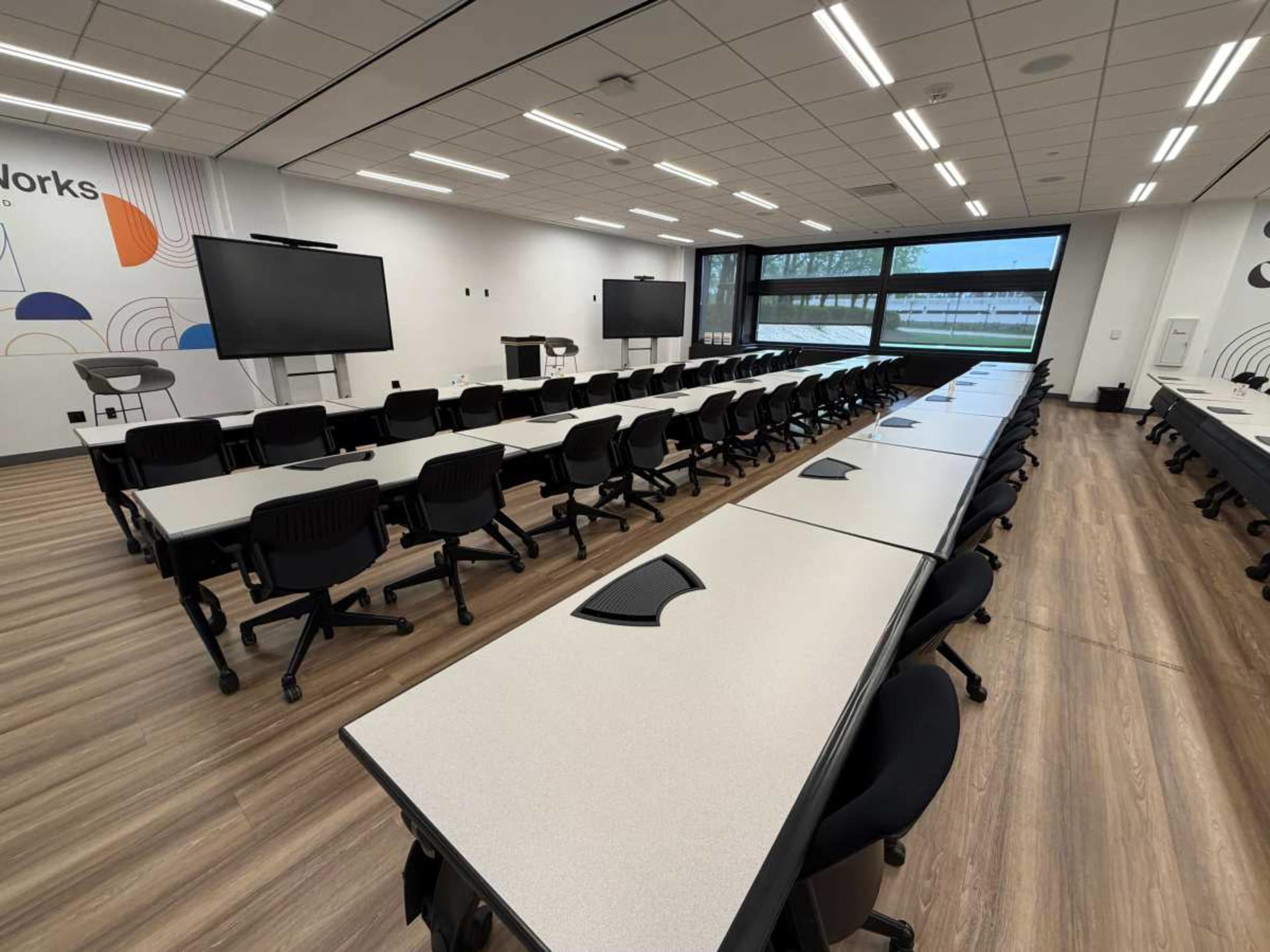 The image shows a modern classroom with rows of tables and chairs arranged for a learning environment, featuring large screens at the front.