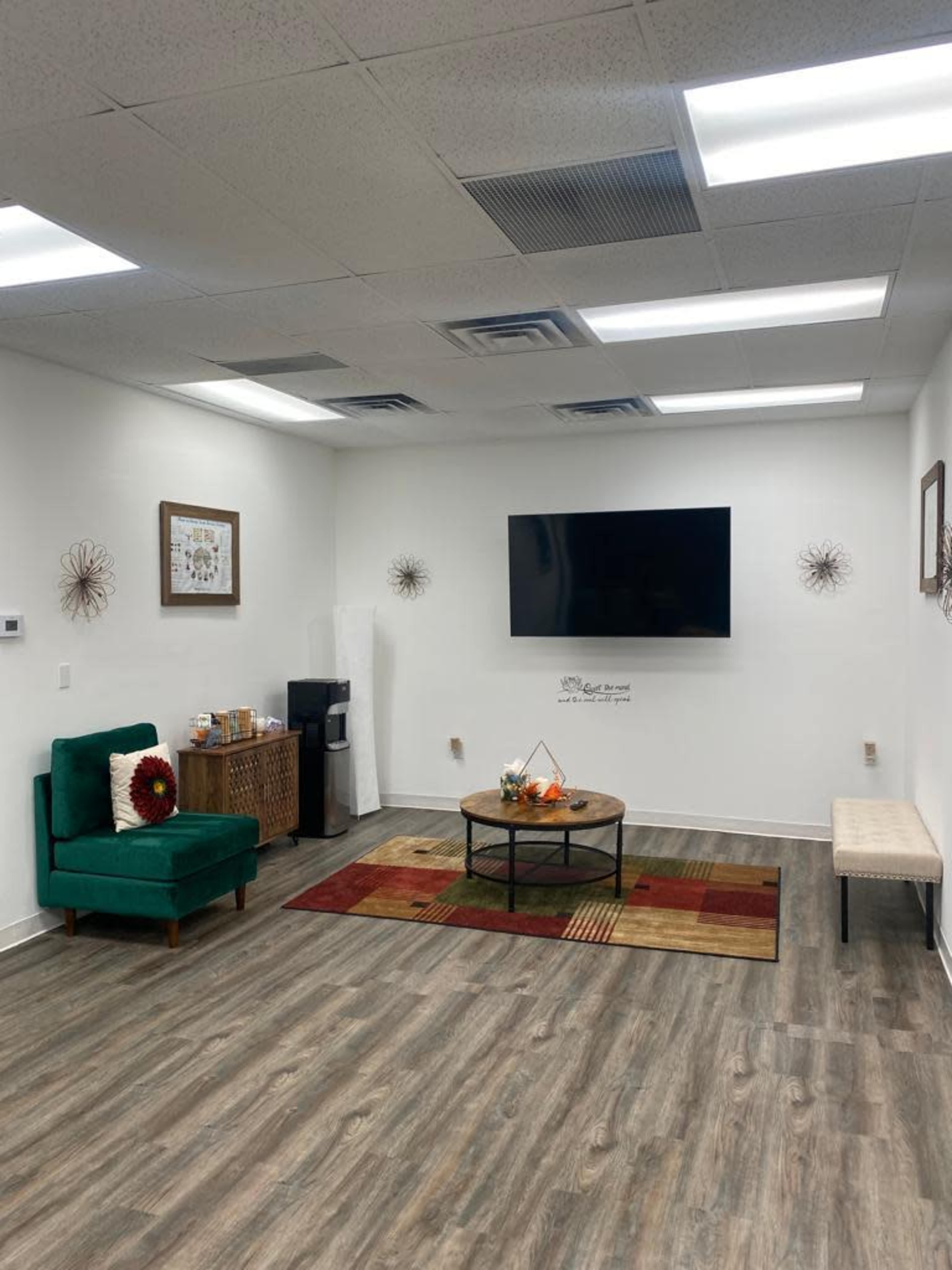 The image shows a well-lit room featuring a green chair, a round coffee table with decorative items, a flat-screen TV on the wall, and a small kitchenette area.