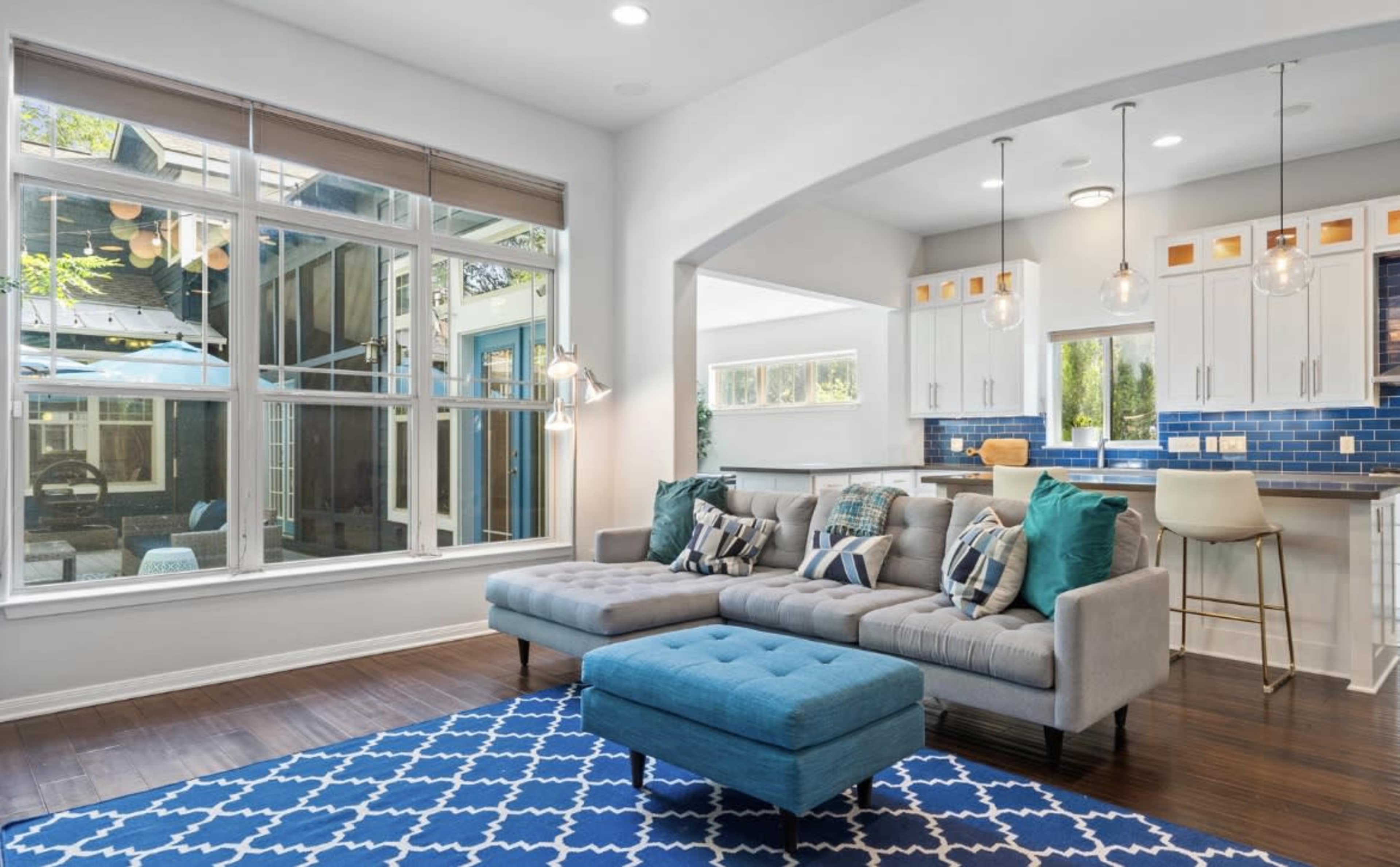A modern living room featuring a sectional sofa, a blue upholstered ottoman, and large windows that provide natural light, with a view of a kitchen in the background.
