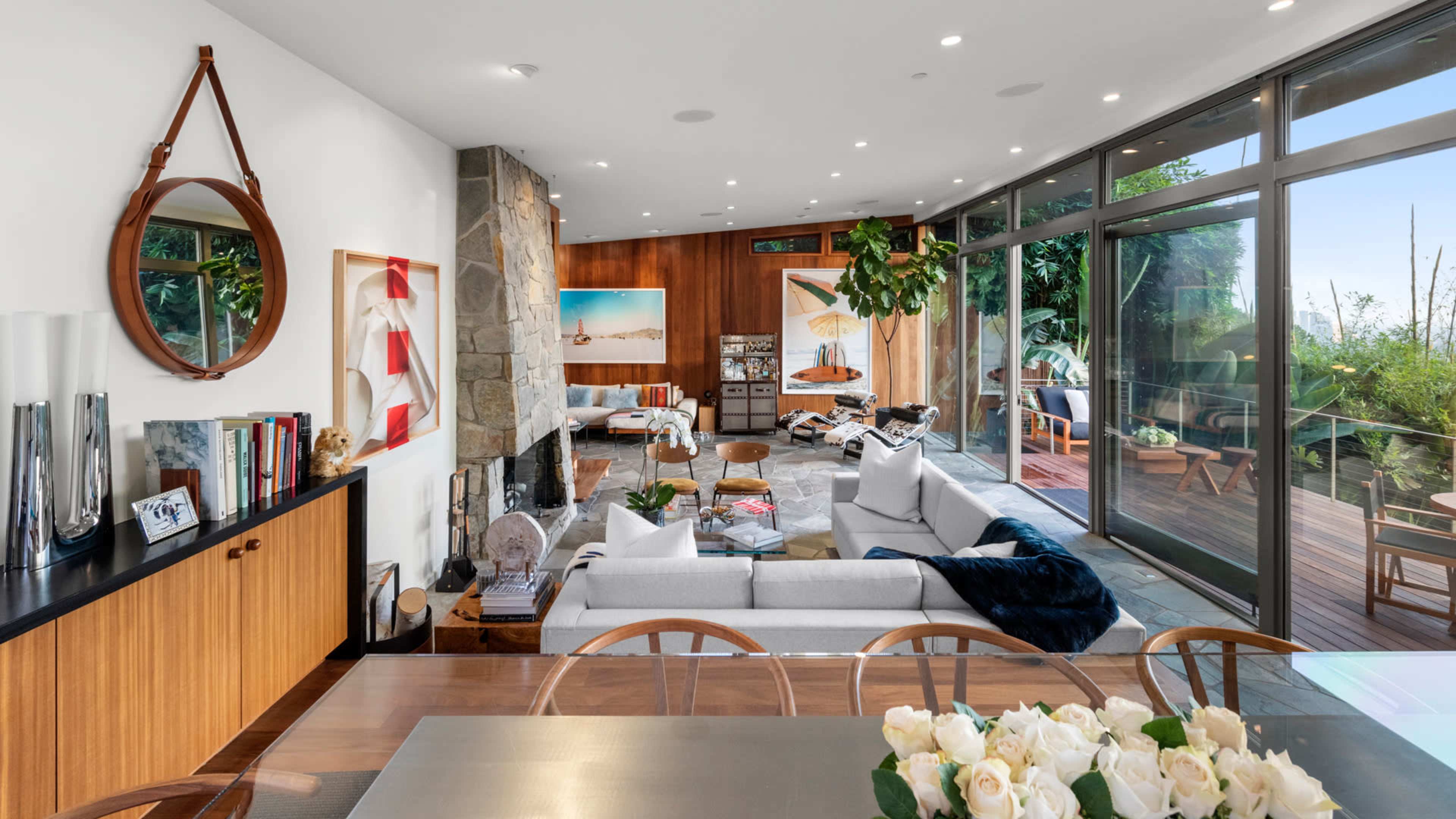The image shows a modern living space with a seamless indoor-outdoor flow, featuring a cozy seating area, large windows, and a dining table adorned with flowers.