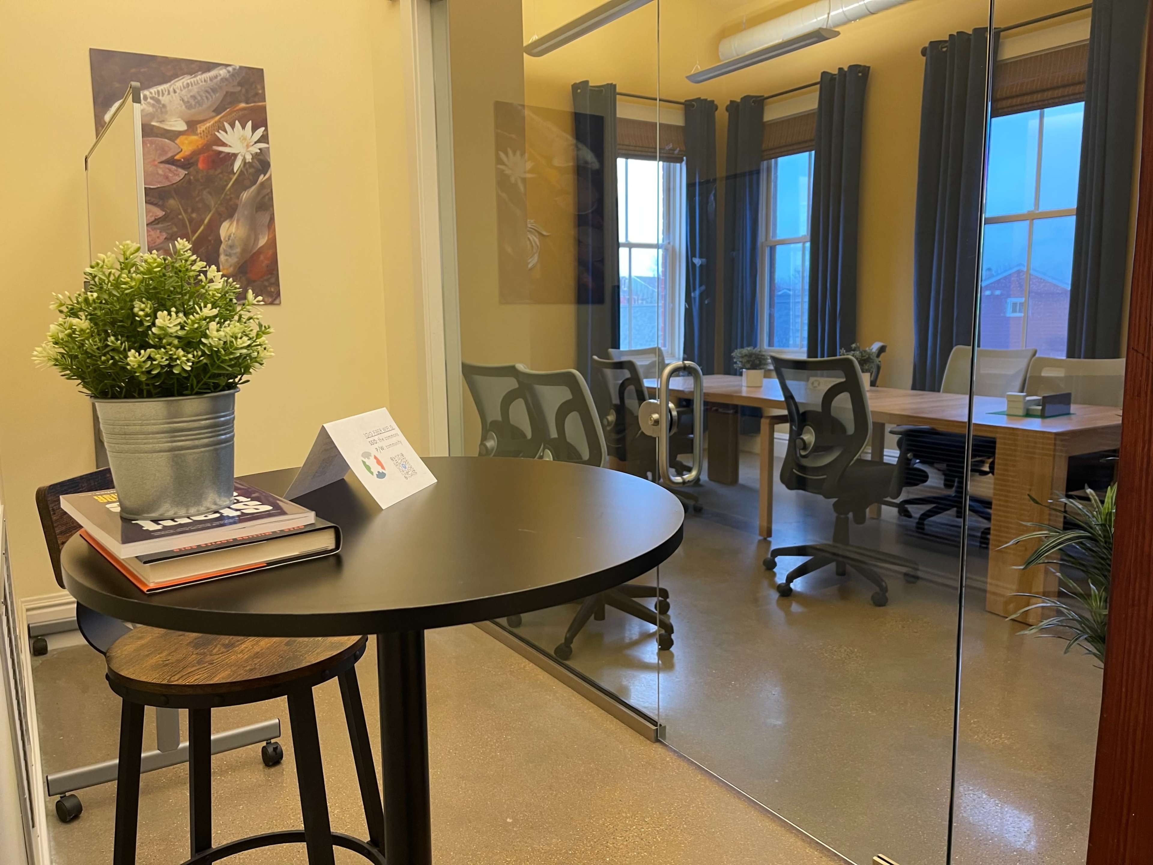 A small table with a plant and books is positioned in front of a glass-walled meeting room containing a large table and several chairs.
