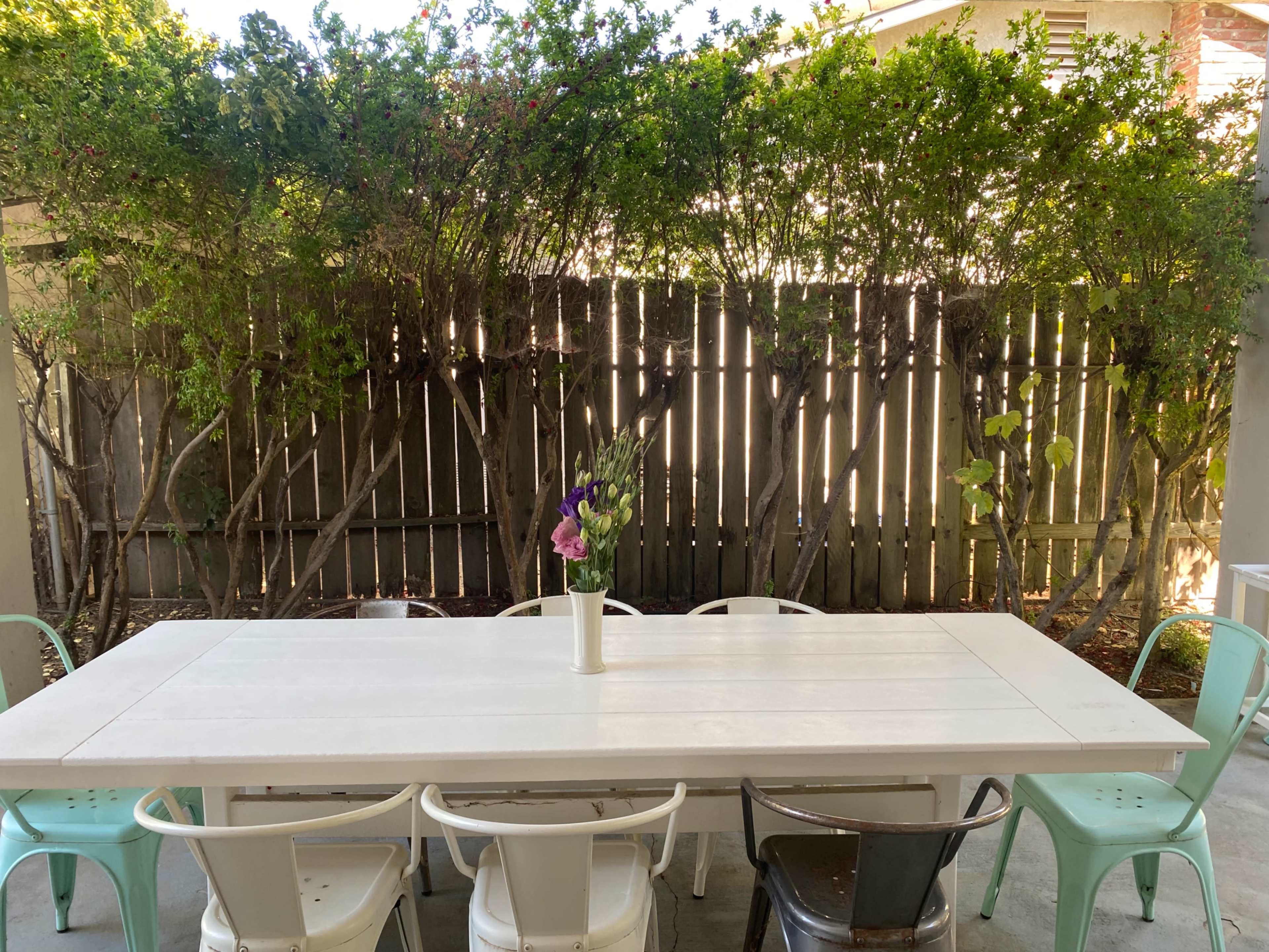 A long white table with a vase of flowers sits in an outdoor space surrounded by a wooden fence and greenery.