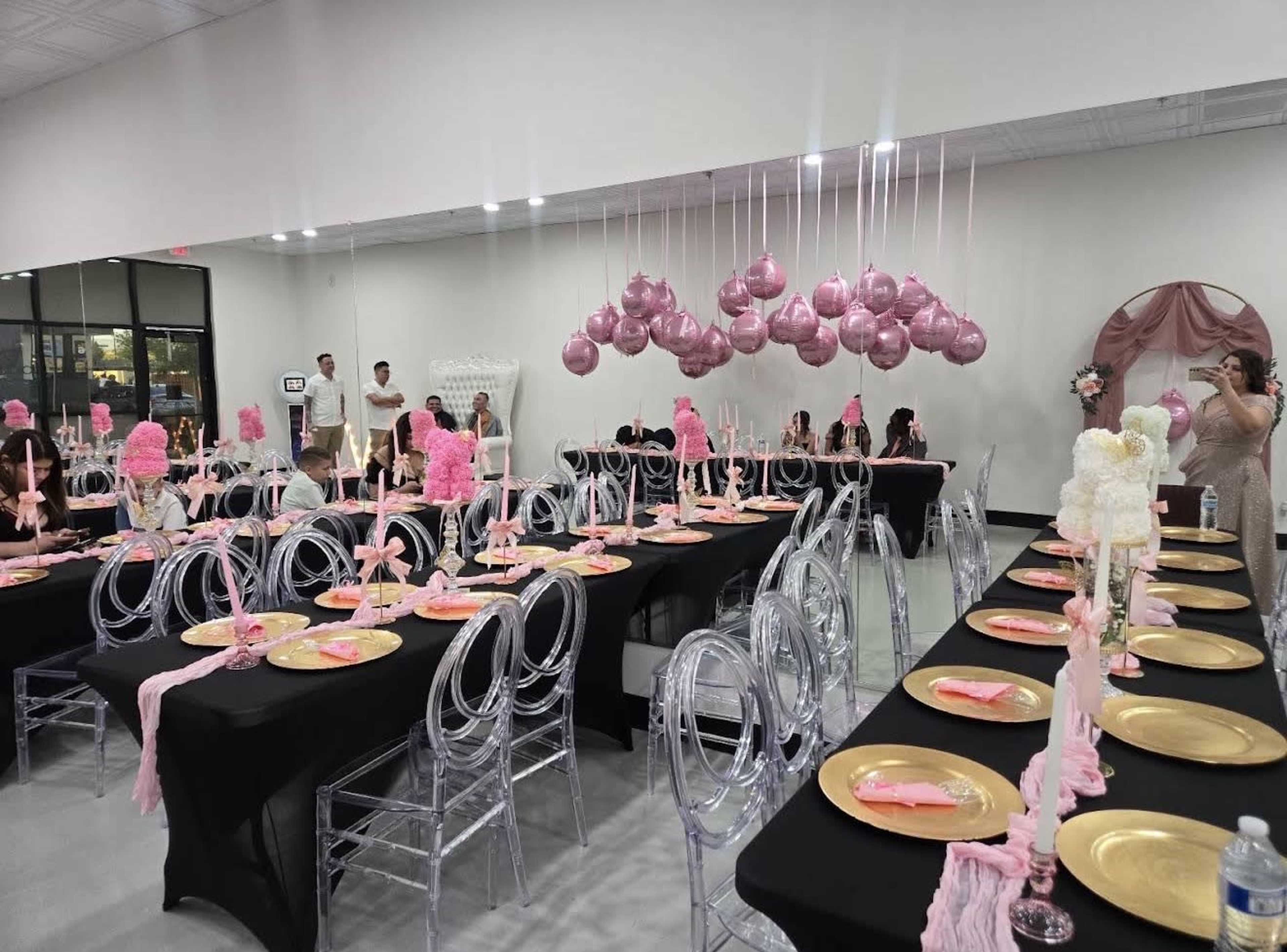 A banquet hall is set with black tables adorned with pink decorations, clear chairs, and gold plates, while pink balloons hang overhead.
