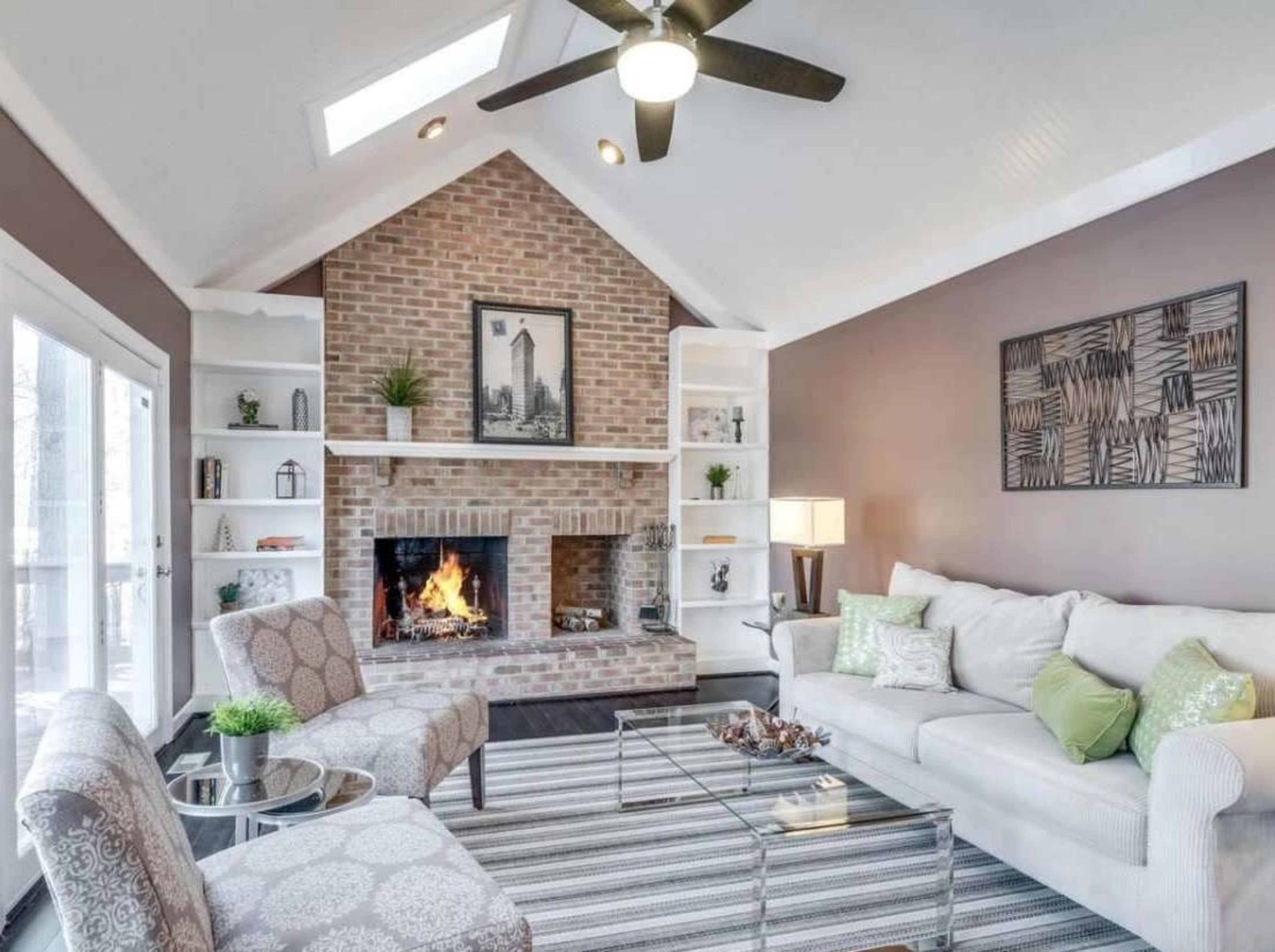 The image shows a cozy living room with a brick fireplace, built-in shelves, and a comfortable seating area featuring a light-colored sofa and patterned chairs.