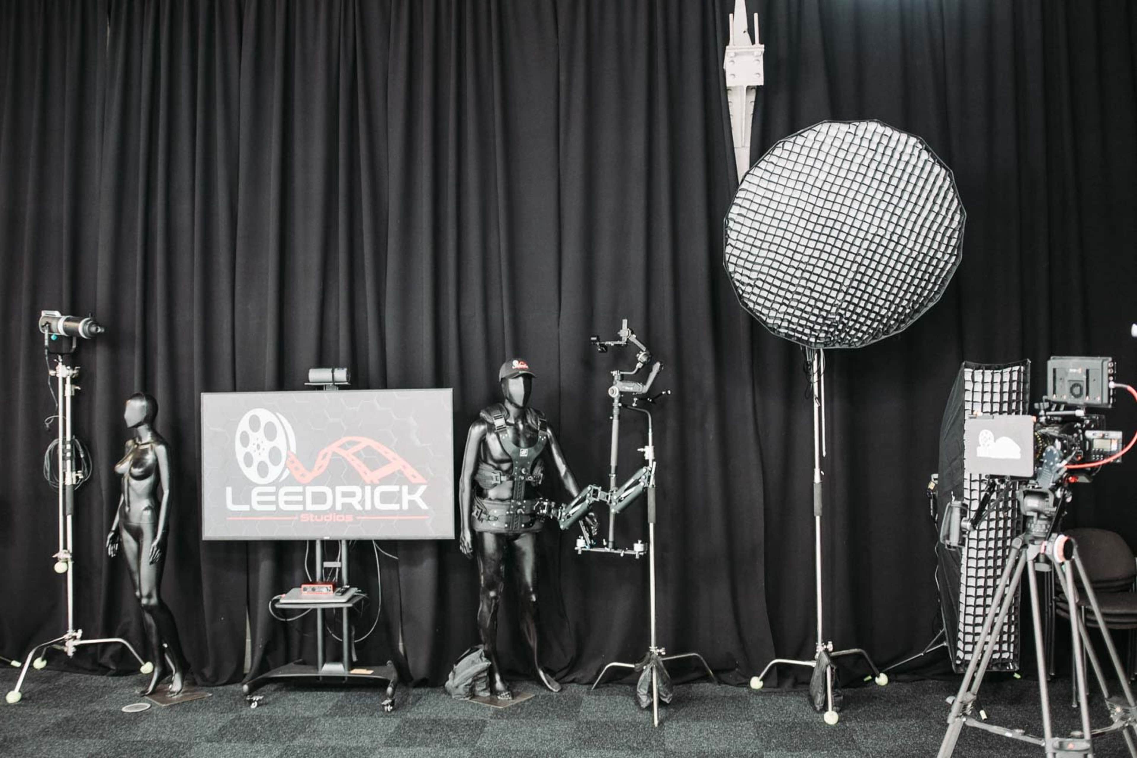 The image shows a photography studio setup featuring two mannequin figures, a large softbox light, cameras on tripods, and a screen displaying the logo for "LEEDRICK."
