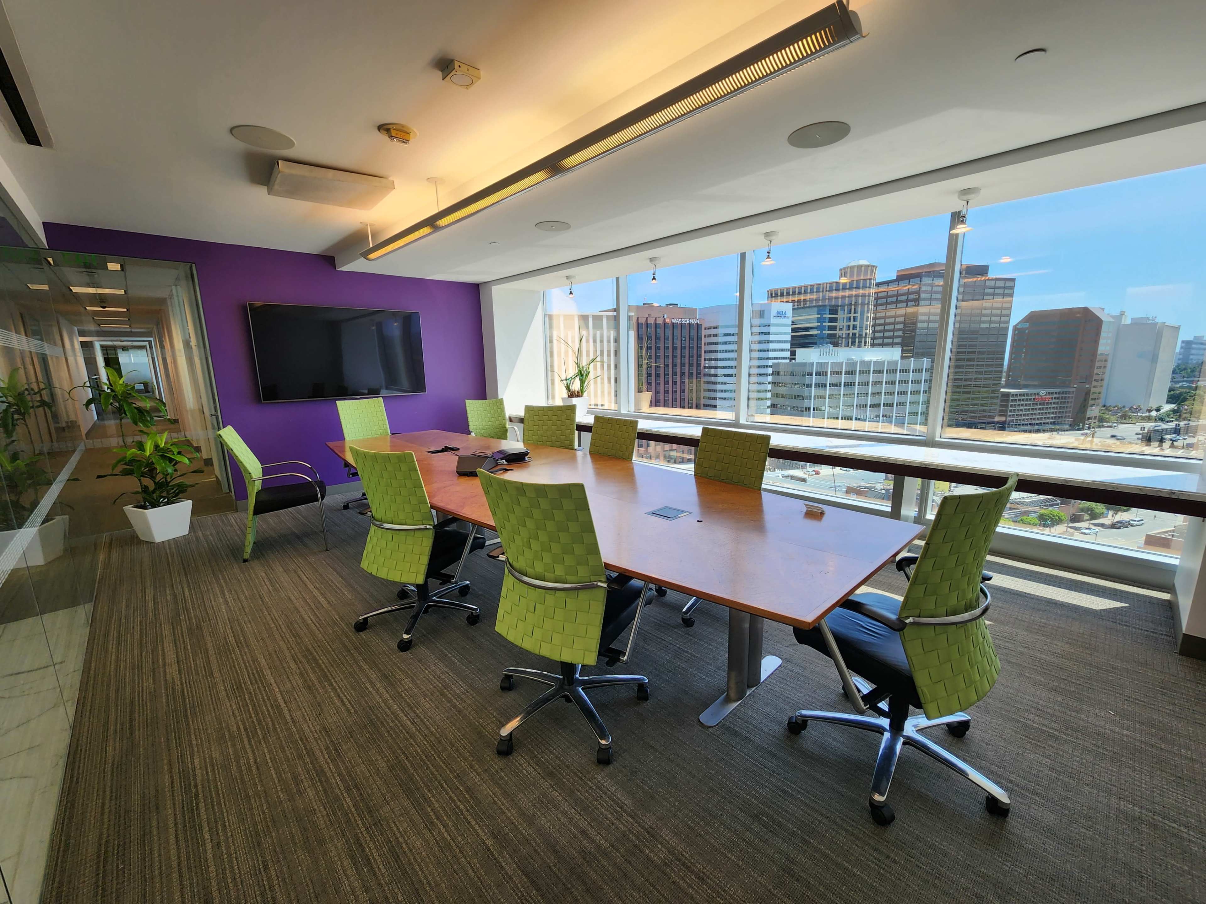 A modern conference room features a large wooden table surrounded by green ergonomic chairs, with a large window showcasing a city skyline.