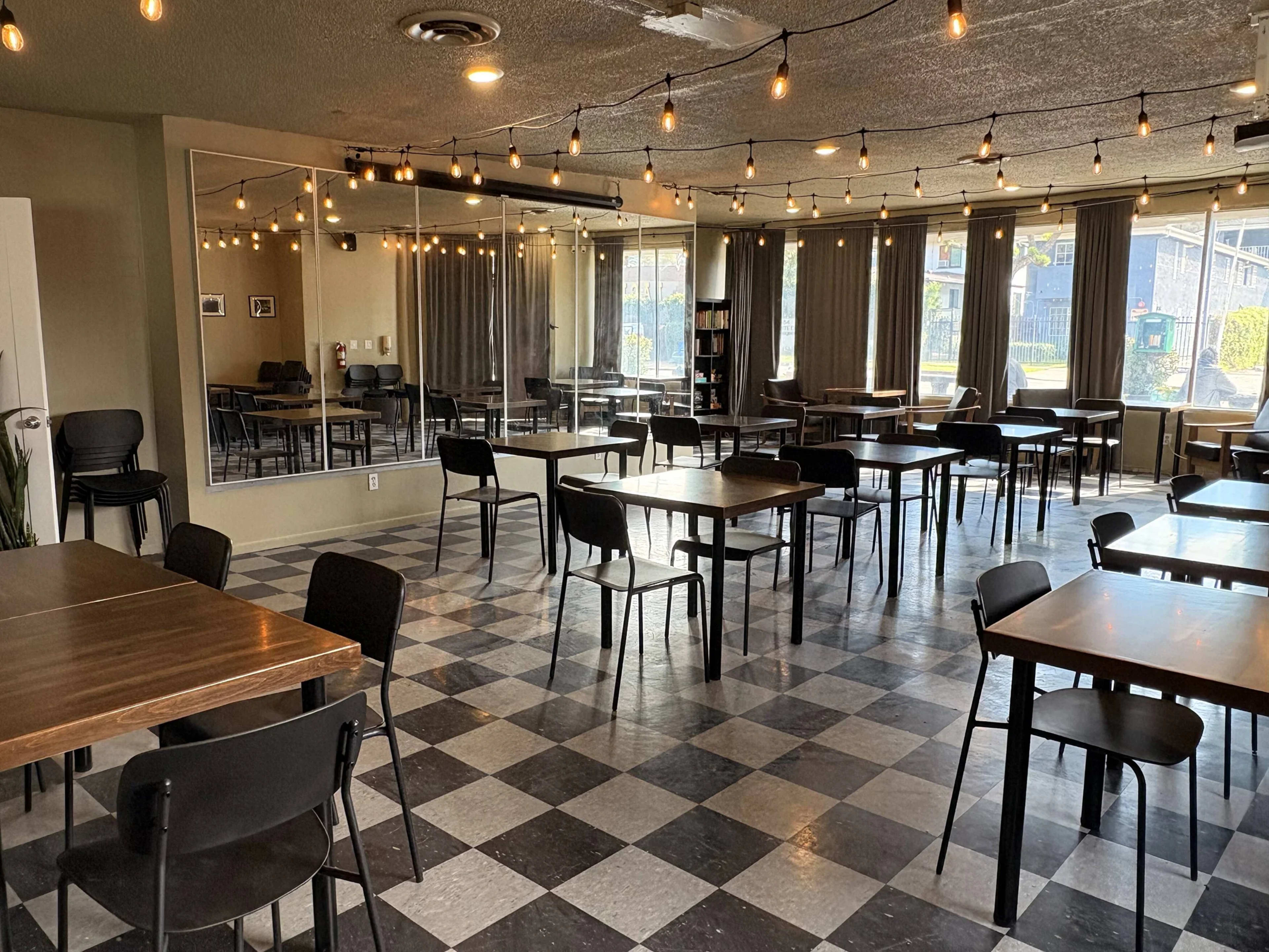 The image shows a spacious, empty dining area with wooden tables and black chairs, featuring a checkered floor and string lights overhead.