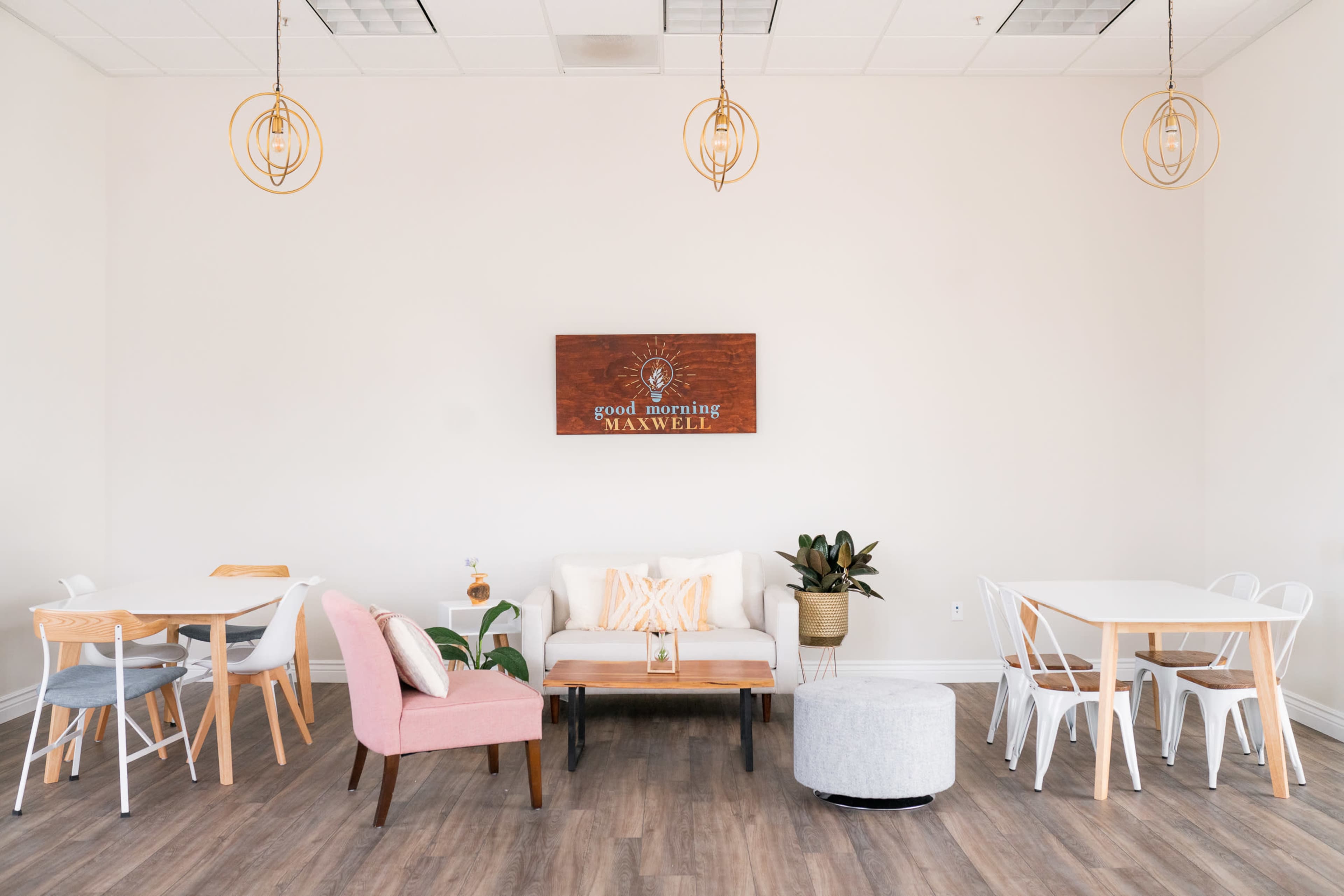 A bright, minimalist room with a mix of seating options, including a couch, chairs, and tables, along with decorative elements like pendant lights and a wall sign.