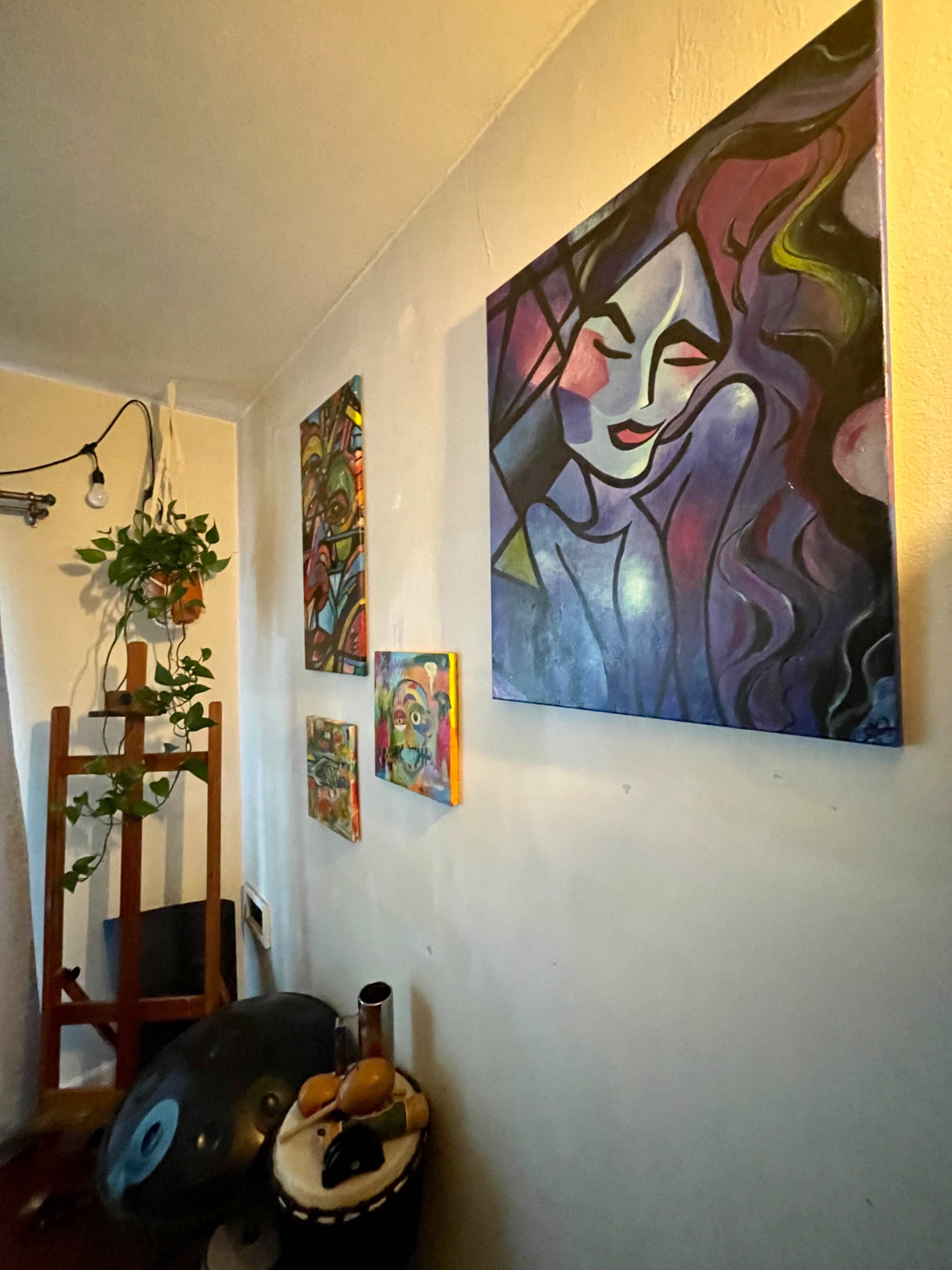 The image shows a wall displaying various colorful artworks, including paintings of abstract figures and a wooden easel beside a small drum.