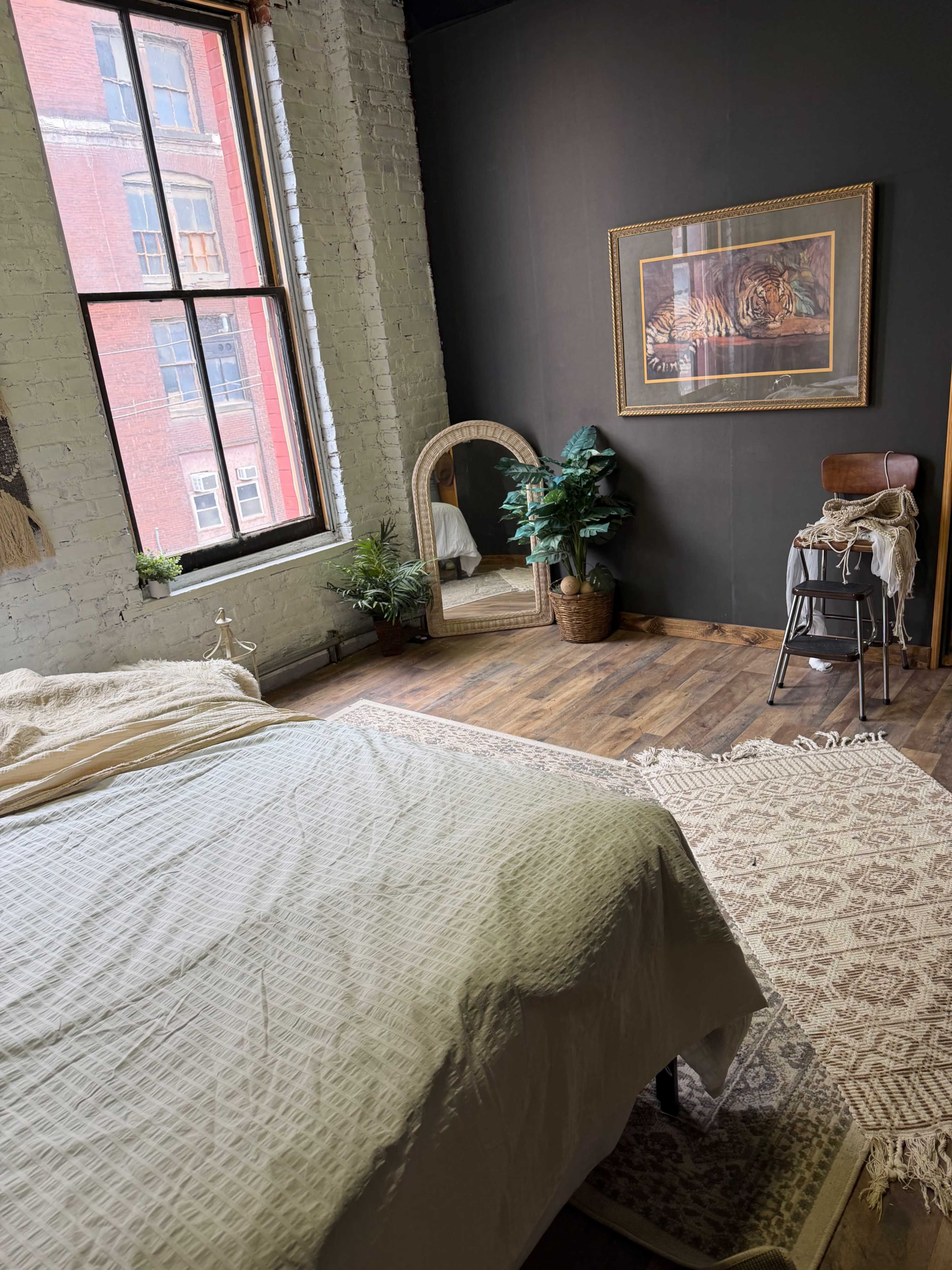 The image shows a cozy bedroom with a large bed, a full-length mirror, a decorative plant, and a wooden chair against a dark wall.