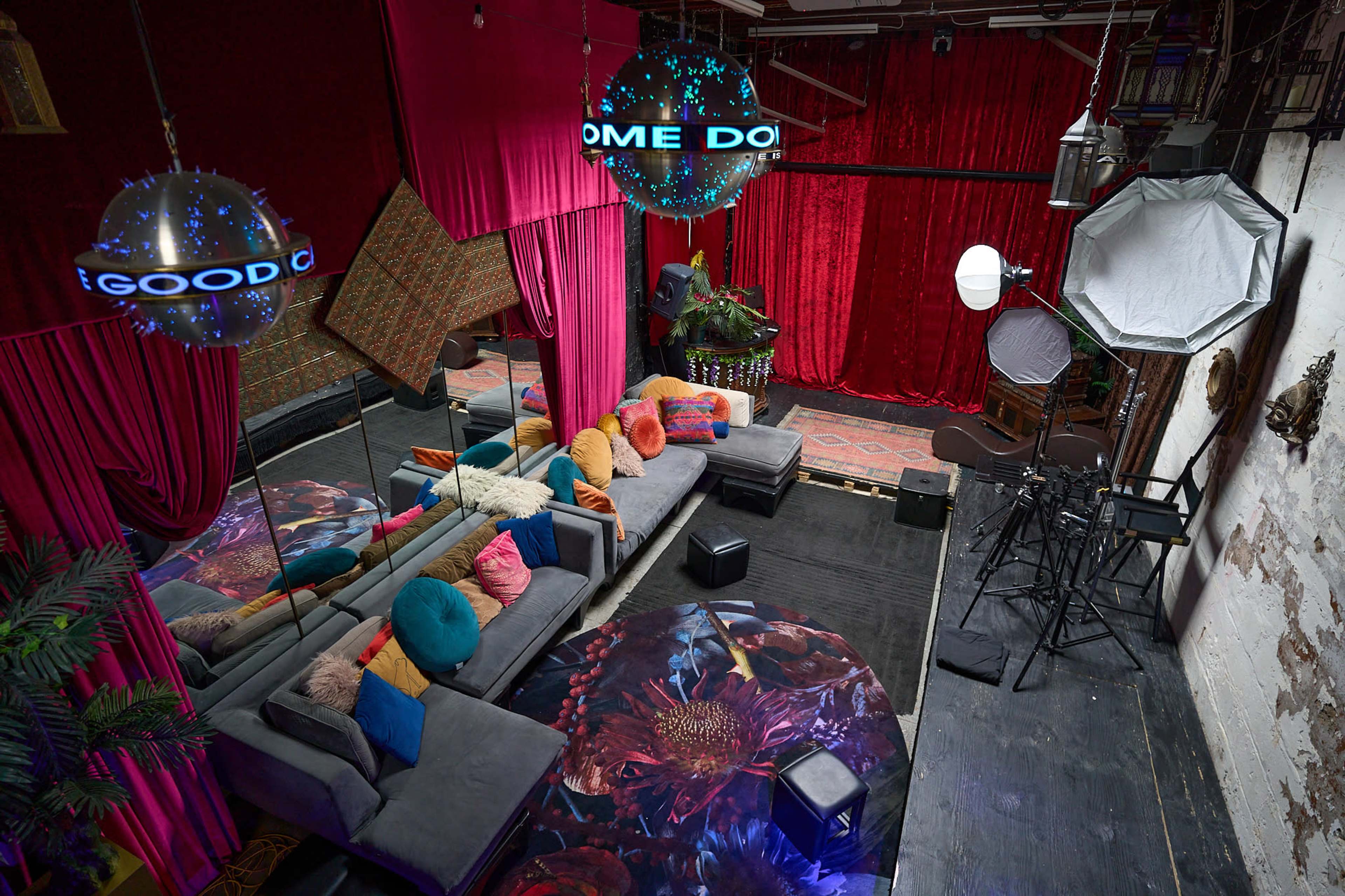 The image shows a vibrant studio space with colorful cushions, patterned carpets, and lighting equipment, surrounded by red velvet curtains.