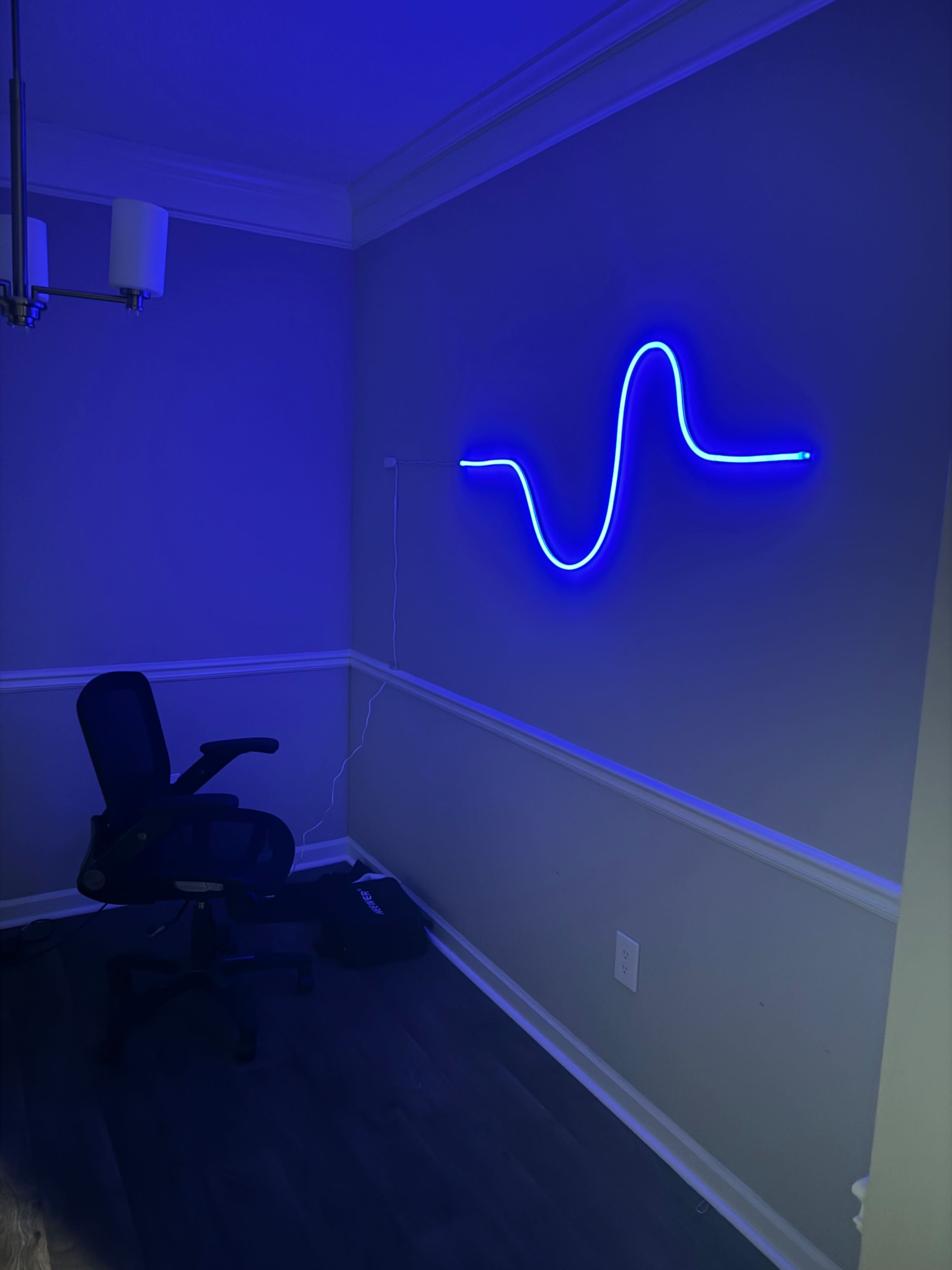 A blue neon light in a wavy pattern is mounted on a gray wall, beside an office chair and dark flooring.