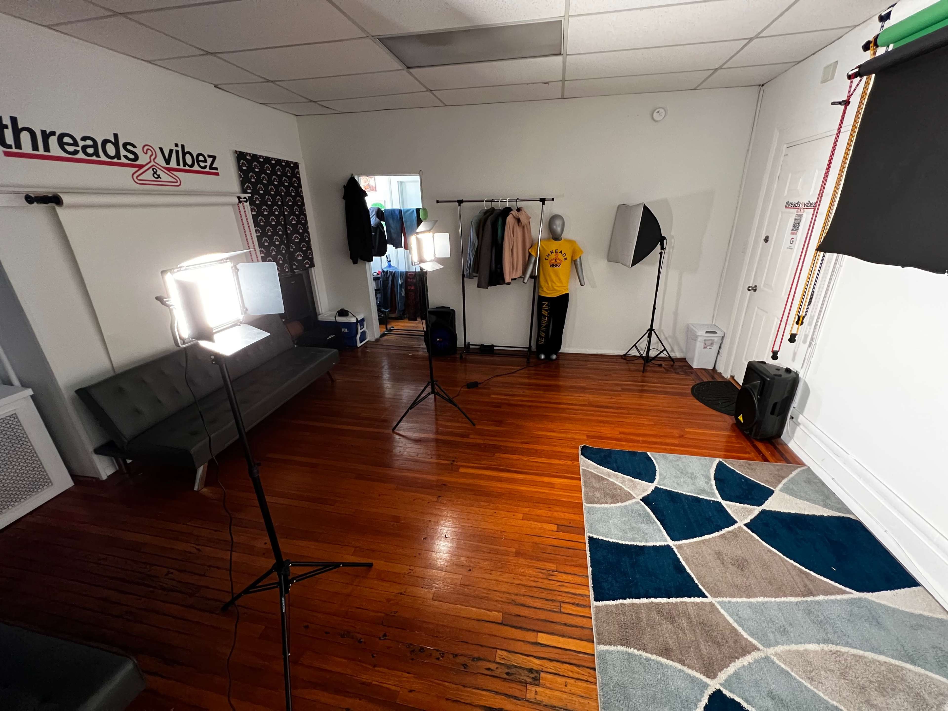 A well-lit room featuring photography equipment, a clothing rack with garments, a sofa, and a patterned rug on the wooden floor.