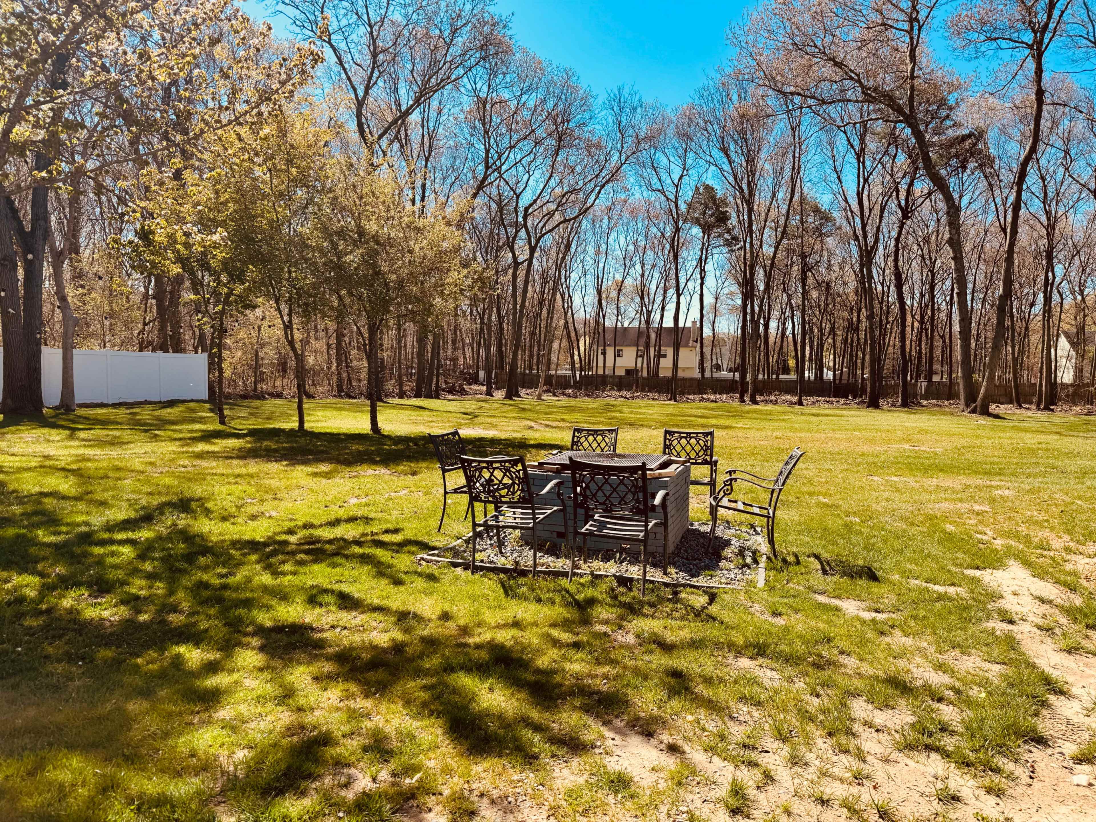 A circular seating area with metal chairs surrounds a stone fire pit, set on a grassy lawn bordered by trees.
