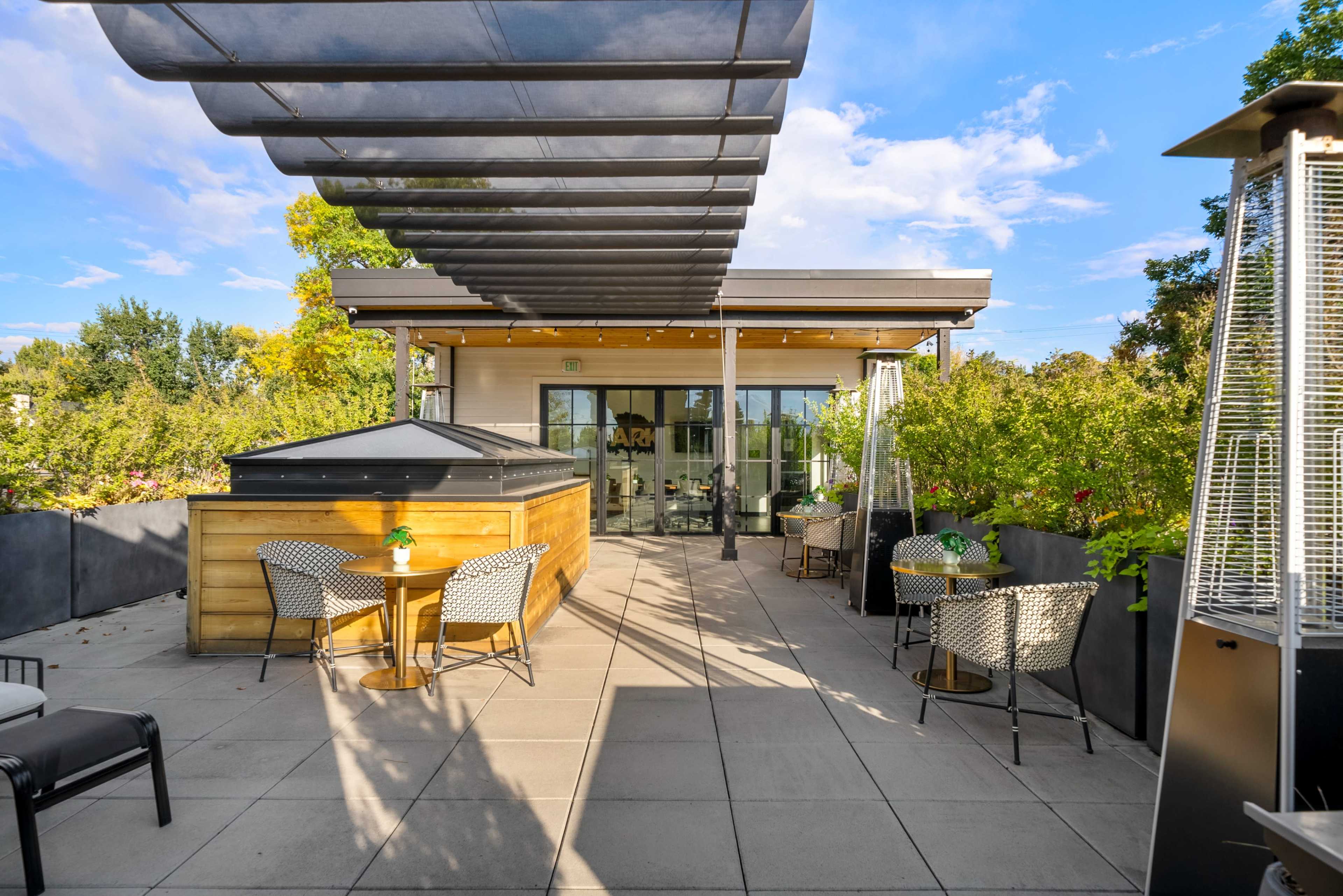 The image shows a modern rooftop patio with seating areas and greenery, featuring a wooden structure and shades overhead.