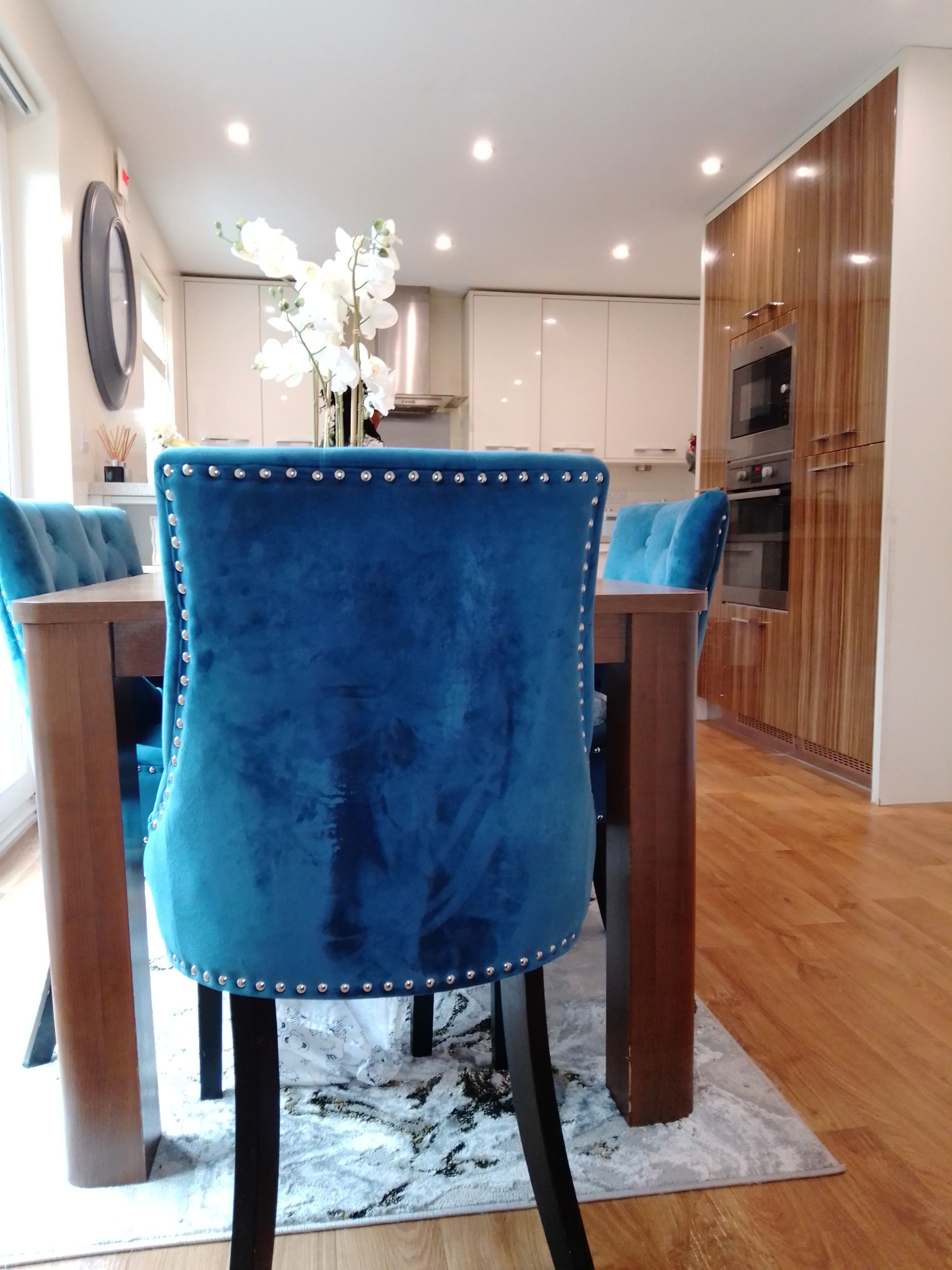 A dining area features a wooden table surrounded by blue velvet chairs with silver embellishments, complemented by a floral centerpiece.