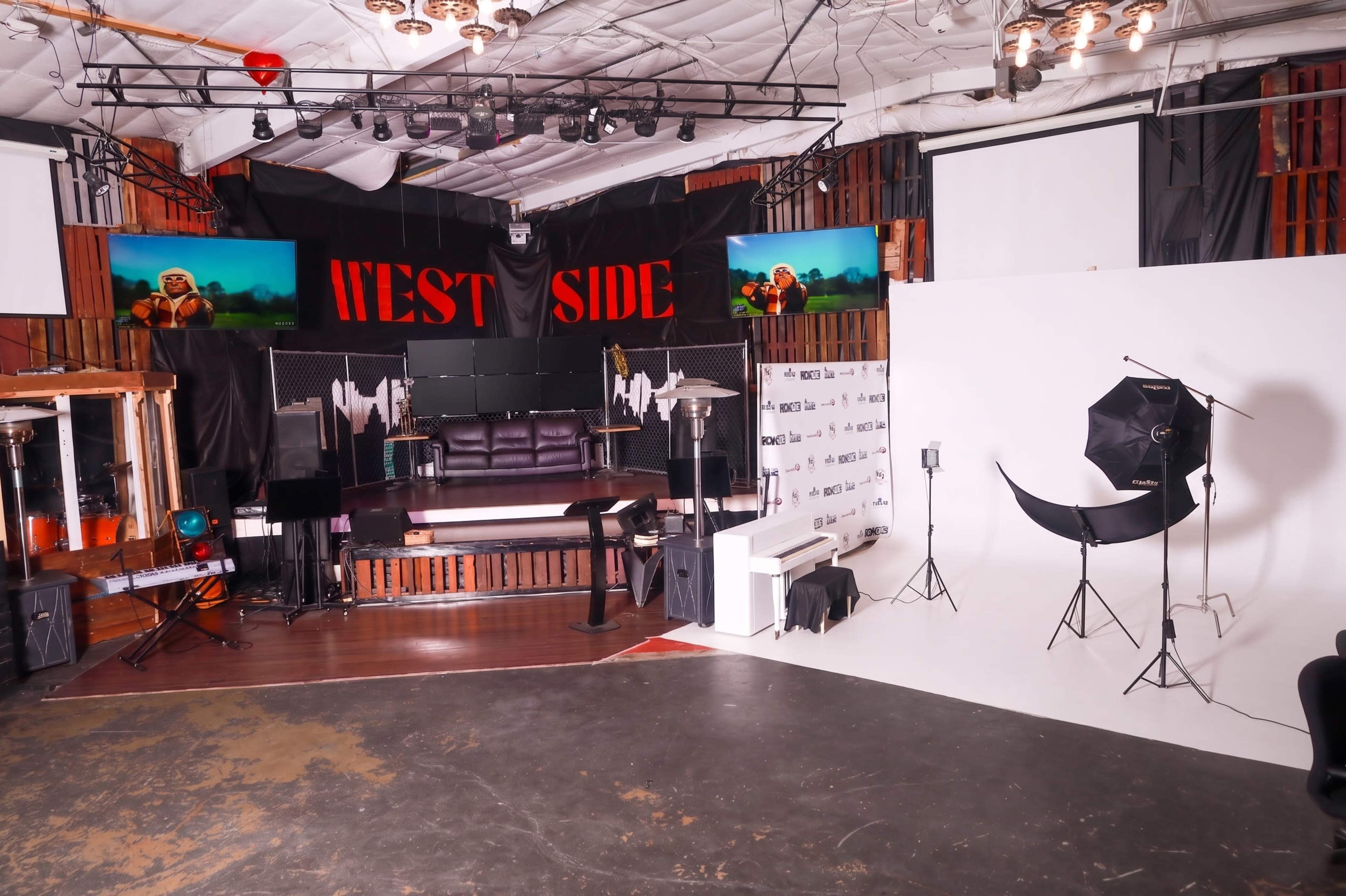 The image shows a multi-purpose studio space with a large stage area featuring a black and white backdrop, television screens, and a seating area.