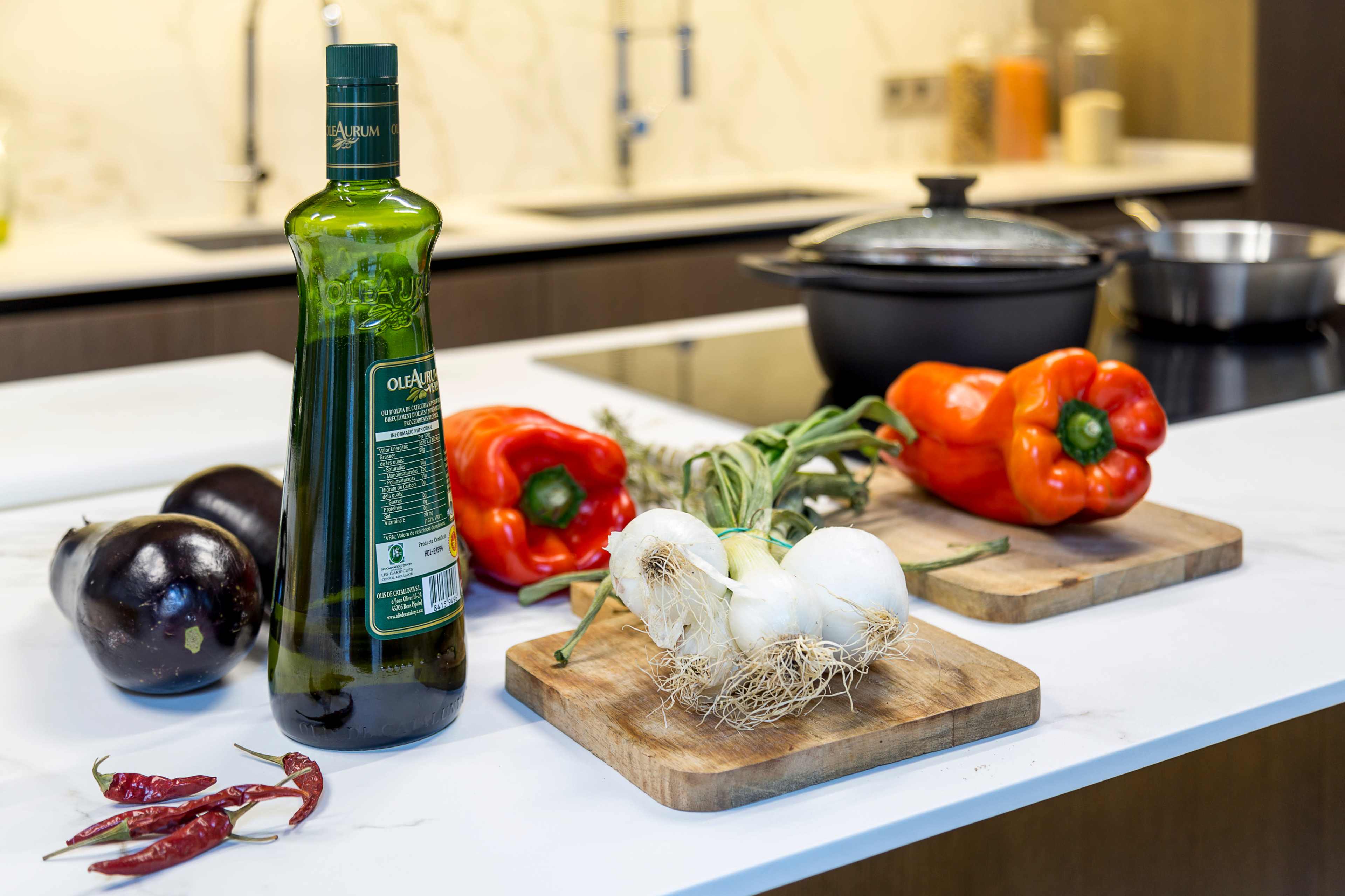 A bottle of olive oil stands next to red peppers, eggplants, and onions on wooden cutting boards in a modern kitchen.