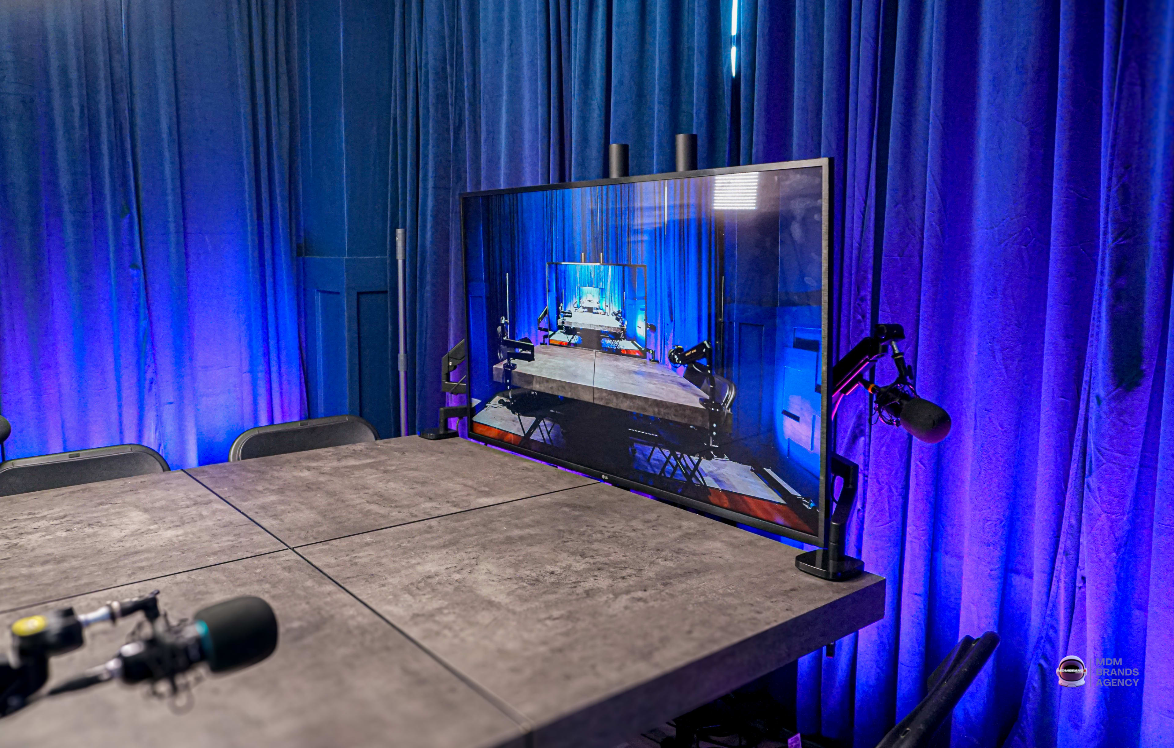 A large screen displays an image of a room with blue curtains and a table, accompanied by microphones, in a dimly lit setting.
