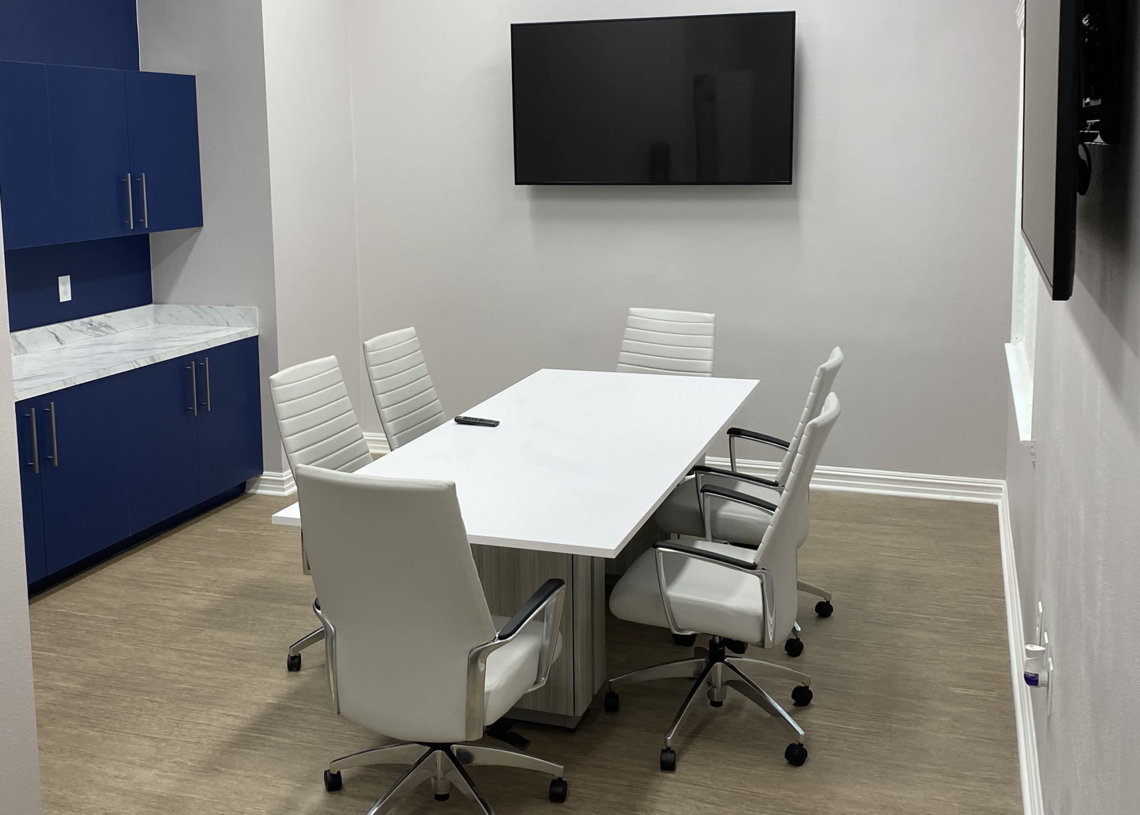 A small conference room featuring a rectangular table with a white surface and five white chairs arranged around it, along with a wall-mounted television and blue cabinetry.