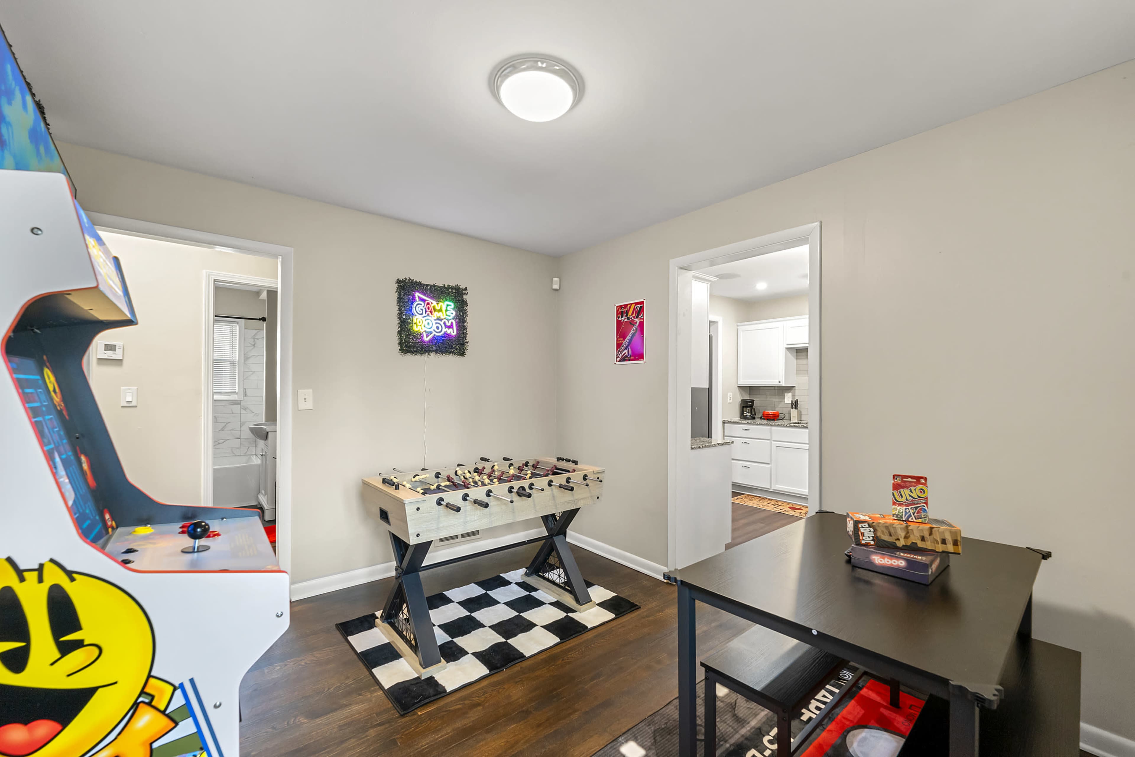 The image shows a game room featuring an arcade machine, a foosball table on a checkered mat, and a small table with a game box, with a doorway leading to a bathroom and a kitchen visible in the background.