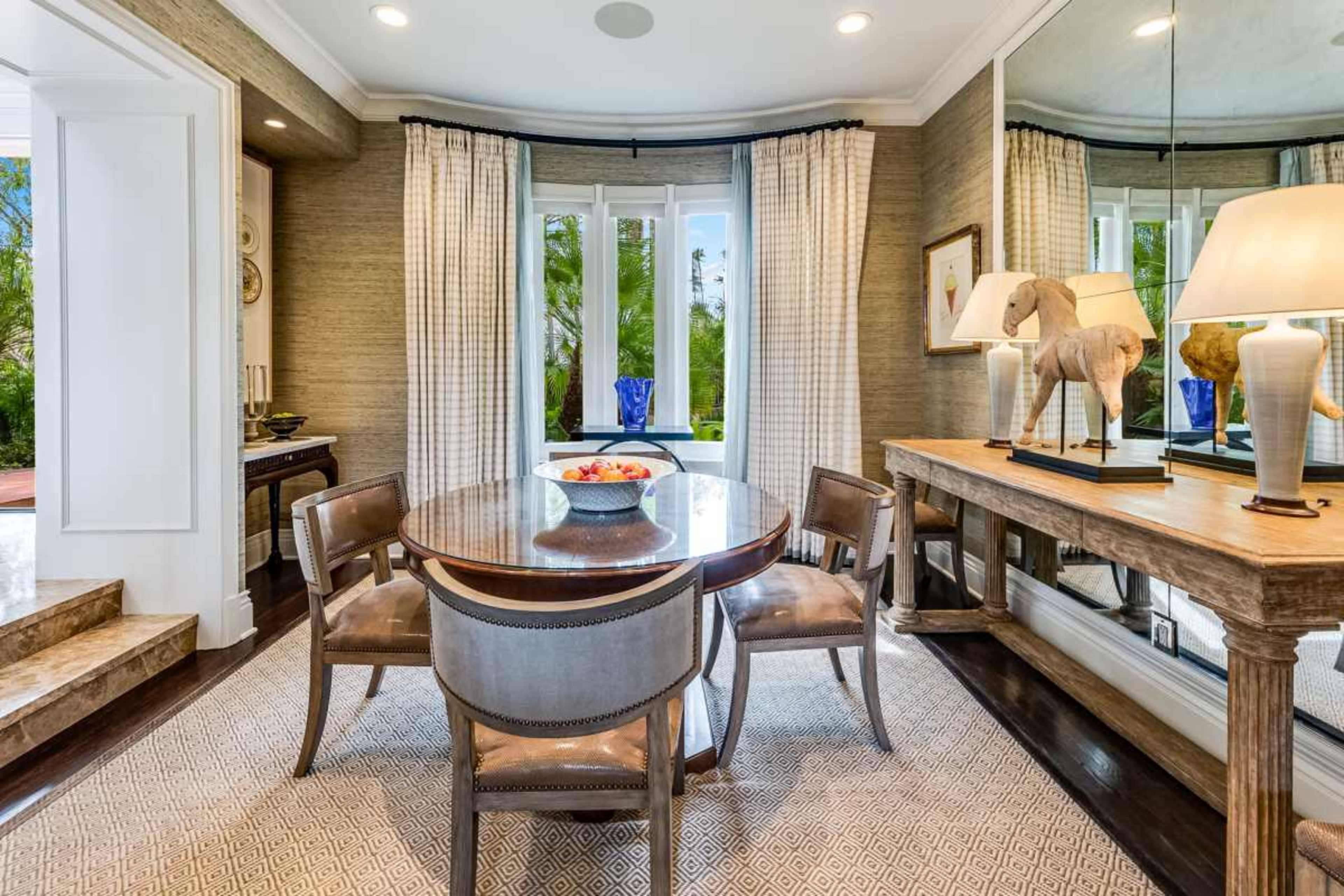 The image features a dining area with a circular table surrounded by four chairs, set against a backdrop of large windows draped with patterned curtains and a decorative mirror.