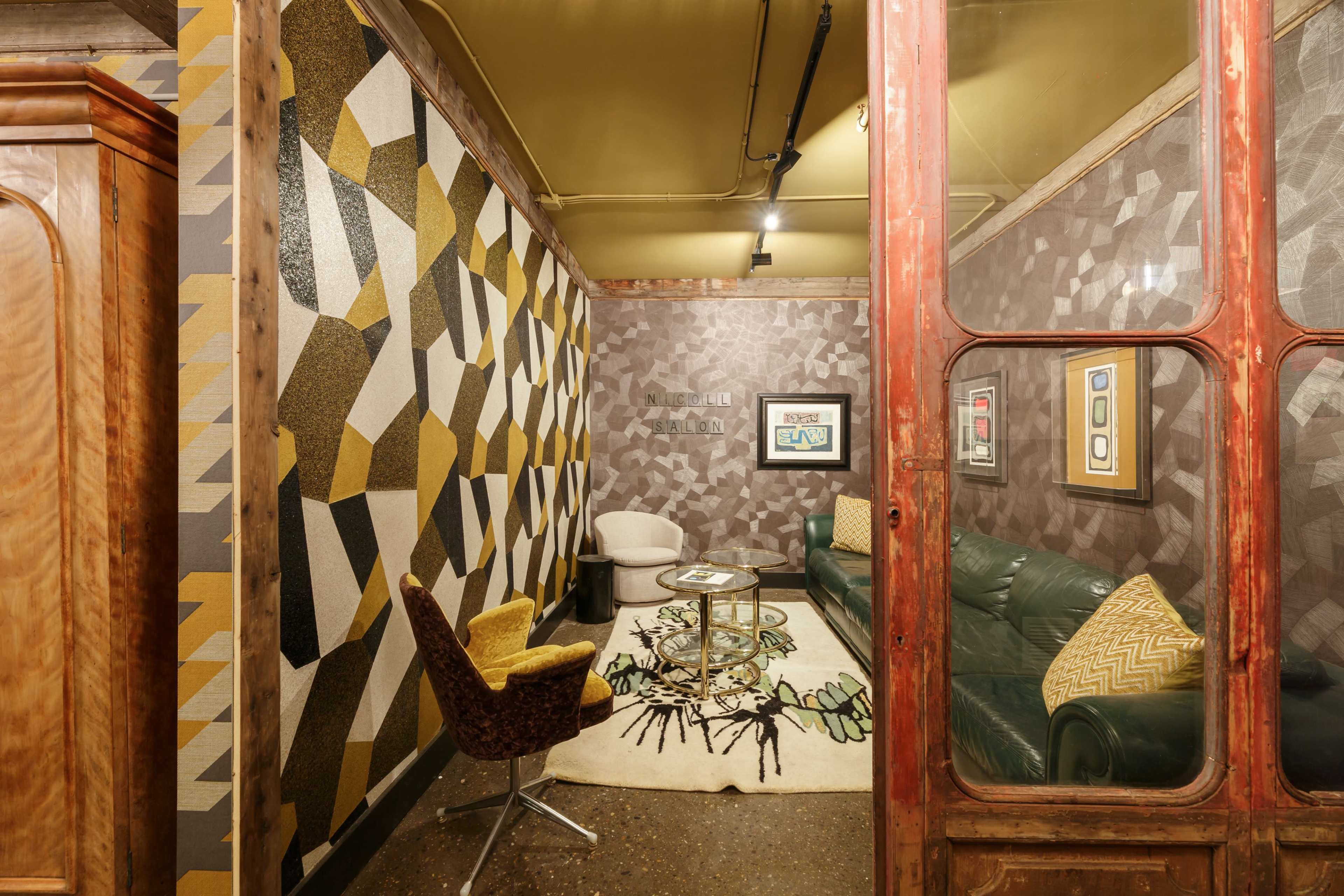 The room features a combination of geometric patterned walls, a green leather sofa, a round coffee table, and a yellow-accented chair, enclosed by a glass door.