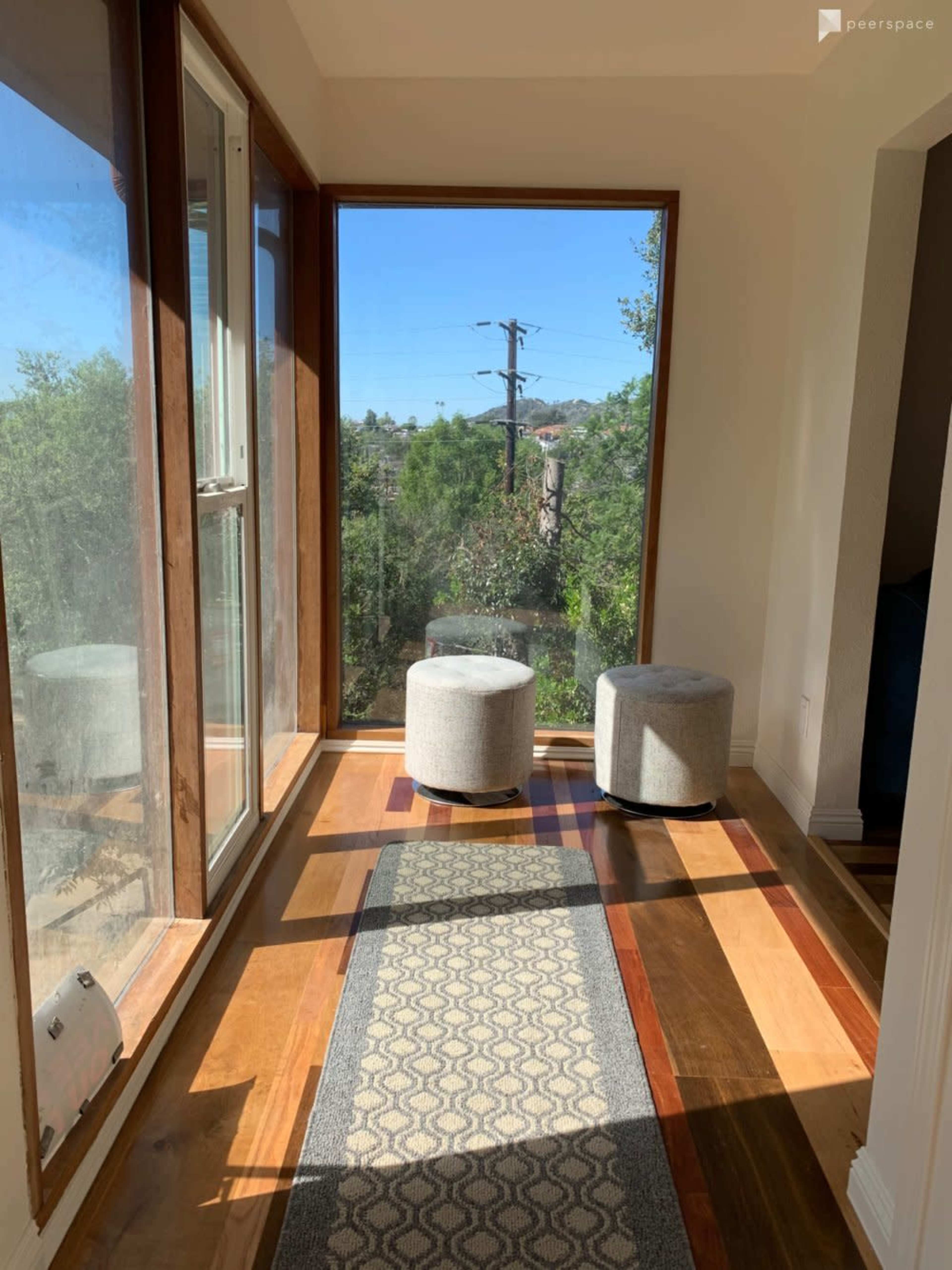 A well-lit hallway features two round stools and a patterned rug beside large windows looking out onto greenery and power lines.