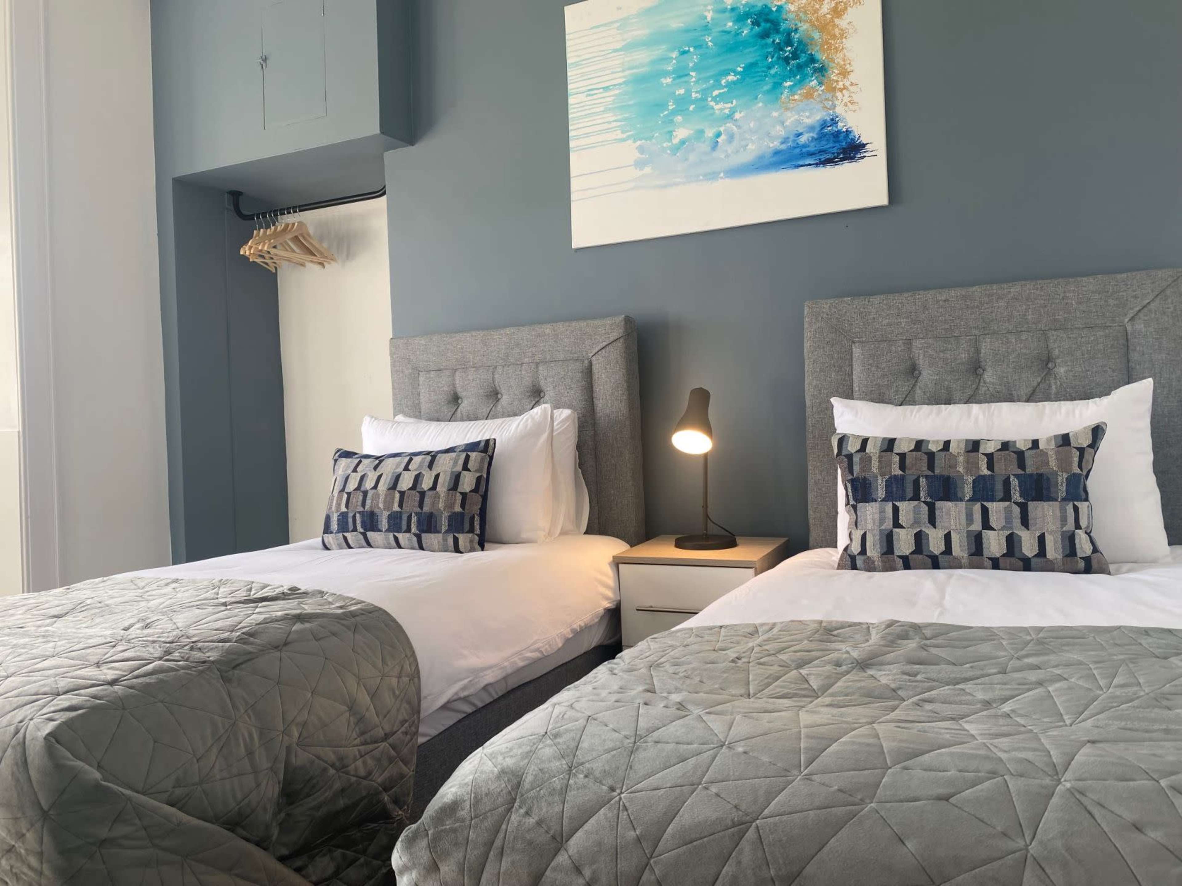 A hotel room with two twin beds, each dressed in white linens and green throws, accompanied by a nightstand and a lamp, against a gray wall featuring an abstract painting.