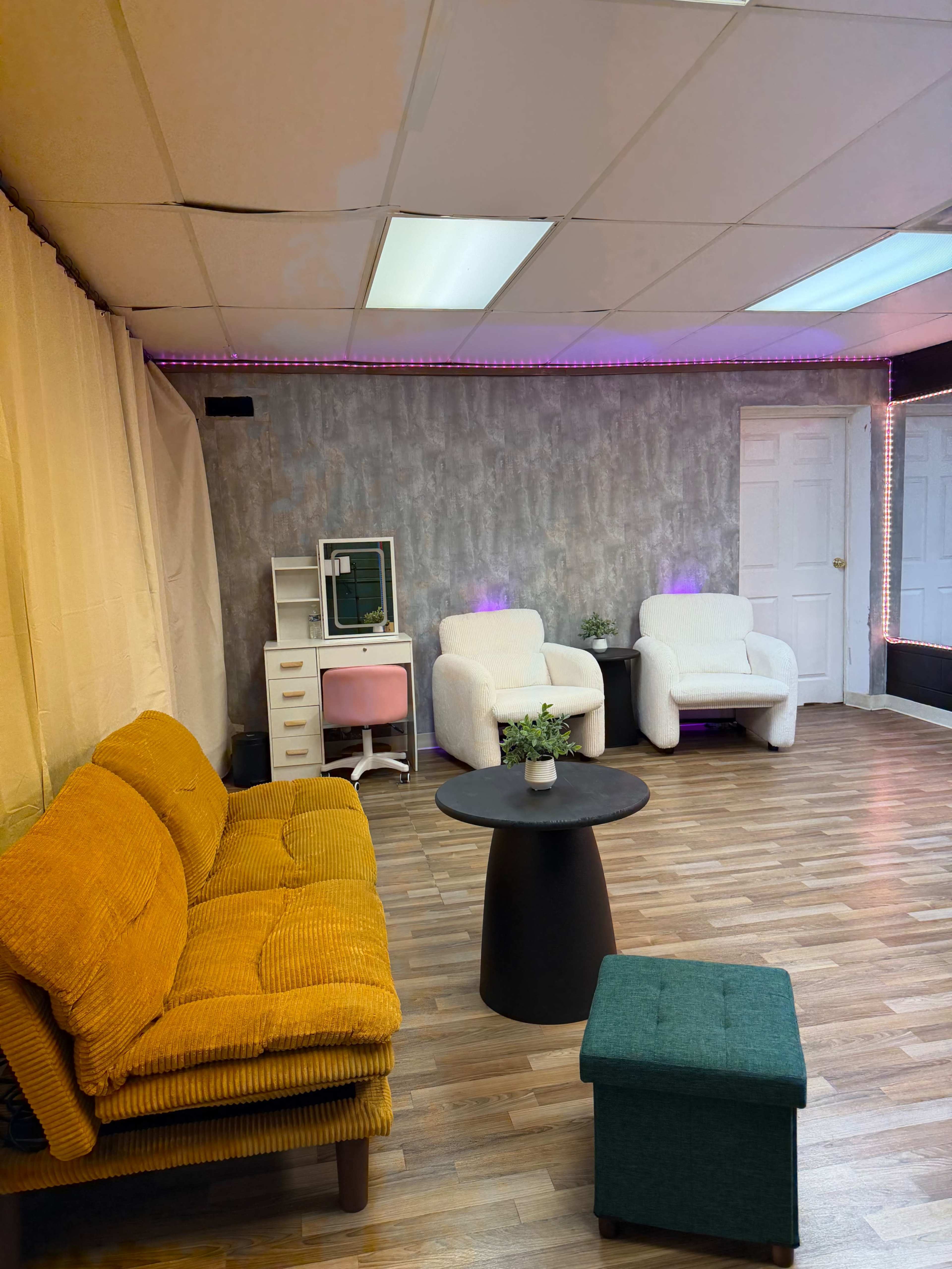 The image shows a modern lounge space featuring a mustard-colored couch, a black circular table, two white armchairs, a green ottoman, and a desk against a textured gray wall with ambient lighting.