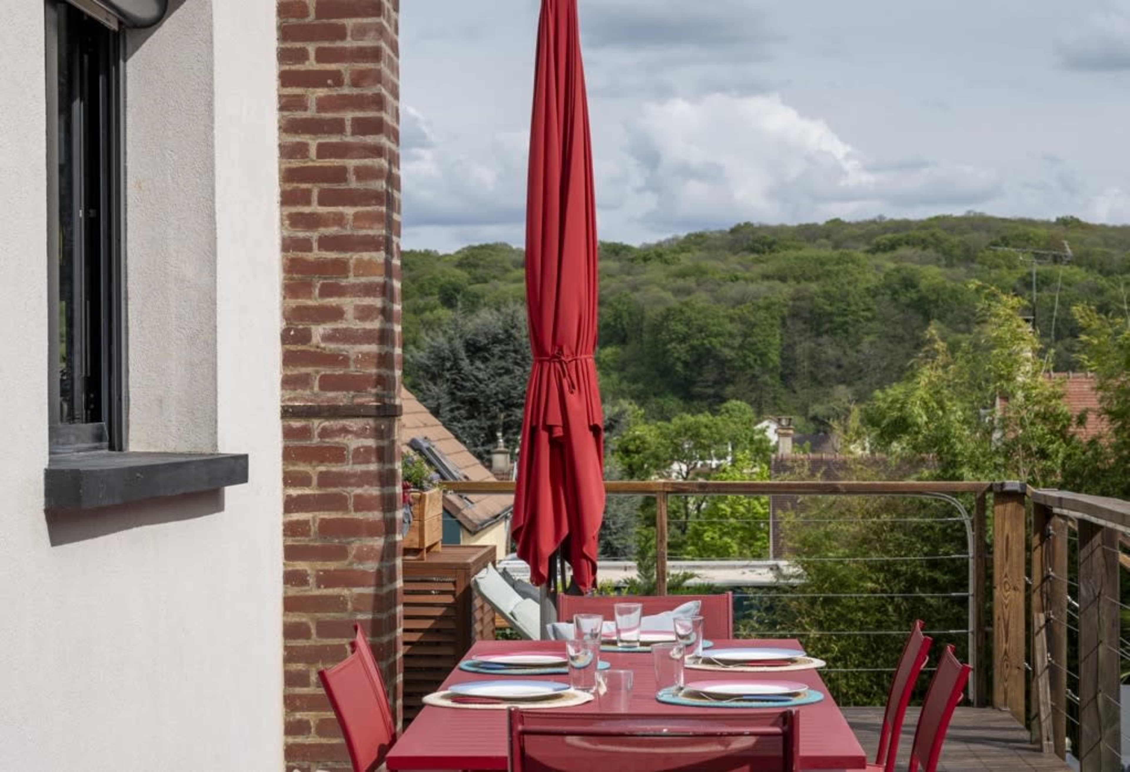 A red patio table set for a meal is positioned on a balcony overlooking a green hillside.