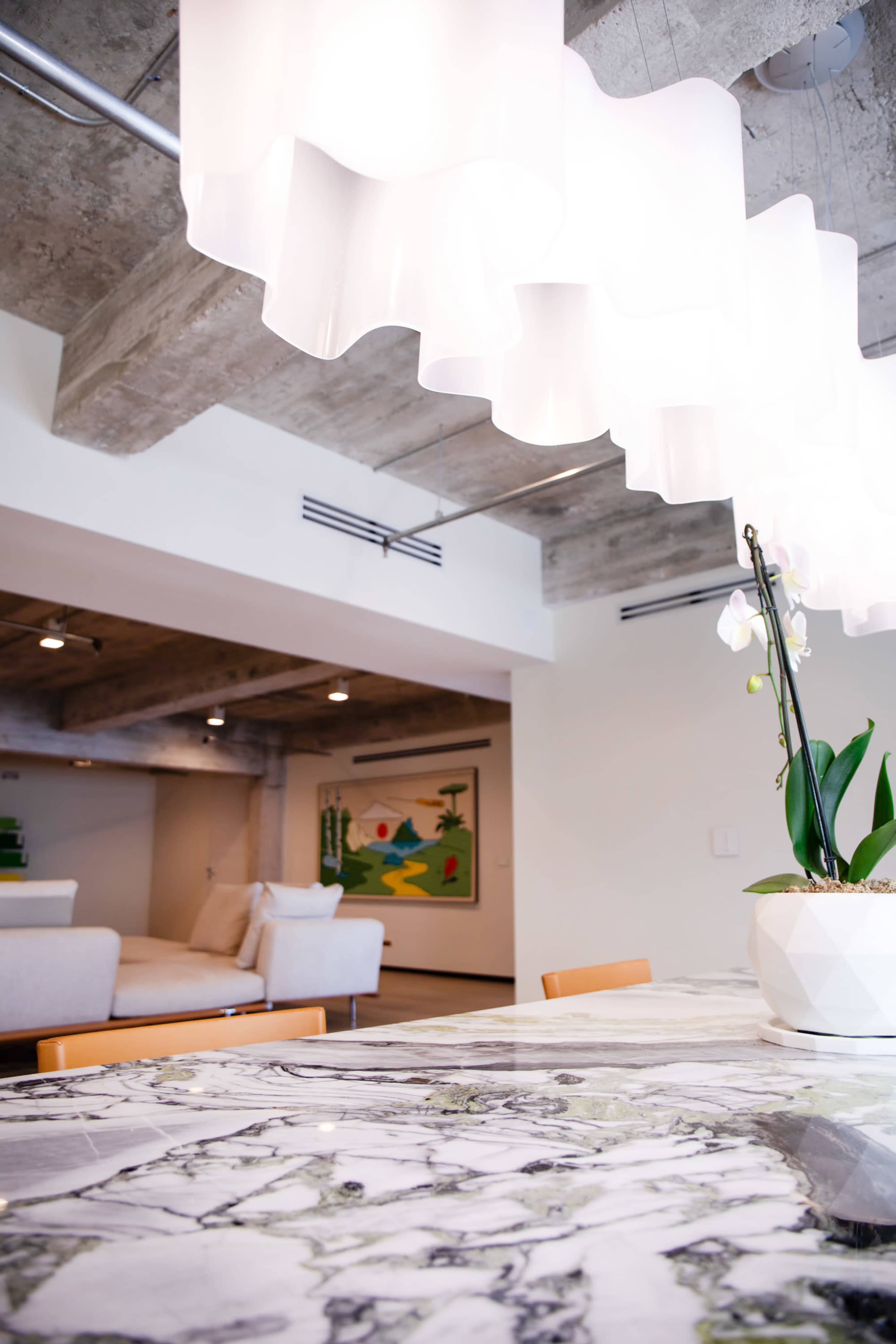 The image features a modern interior space with a dining area, a large marble table, and a decorative white chandelier, leading into a cozy living room.